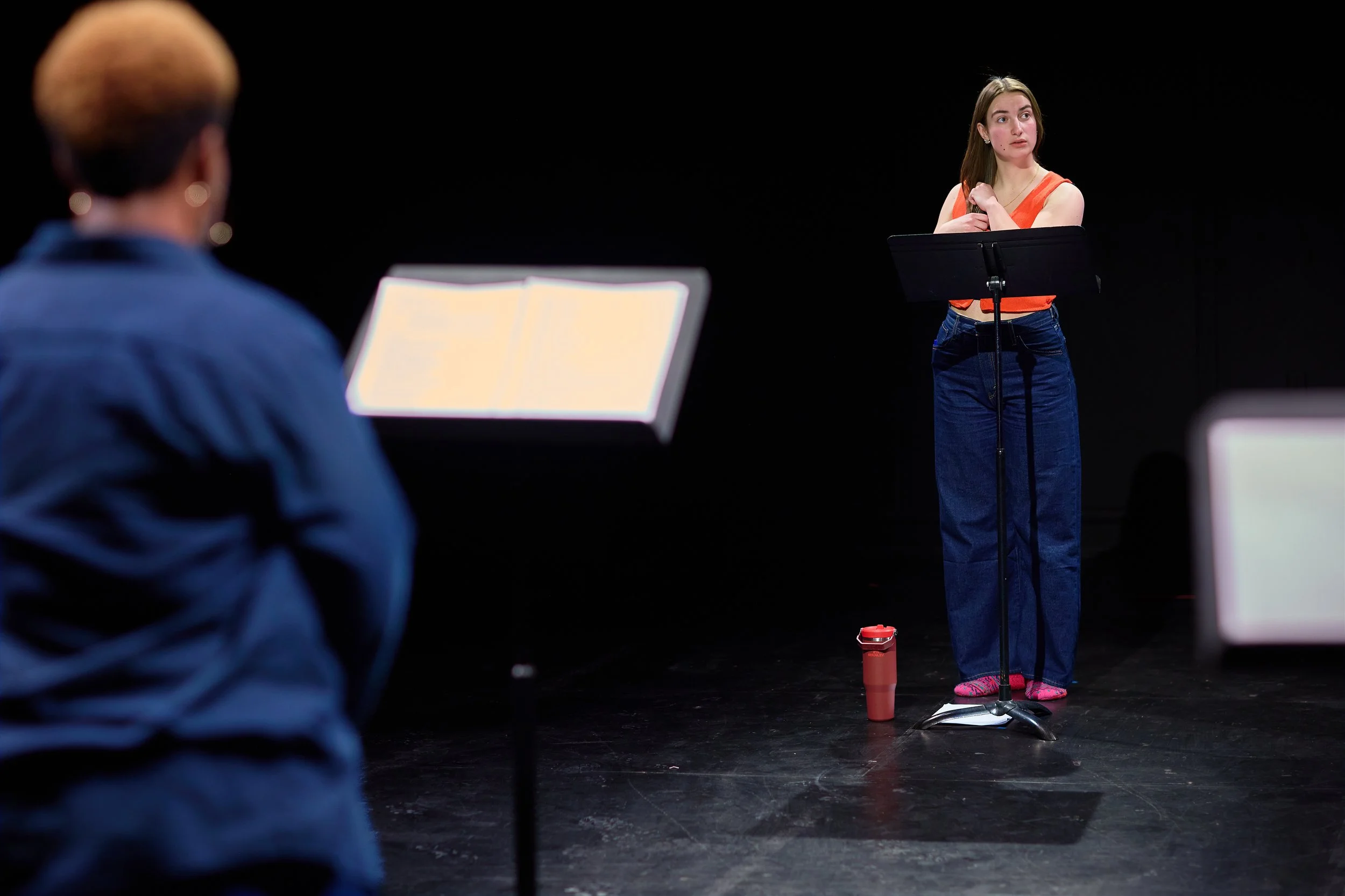 Une jeune femme debout sur scène, en train de parler, avec un pupitre et des partitions devant elle, lors d'une séance de pratique ou de répétition. Deux autres personnes sont en première plan, floues, en face d'elle.
