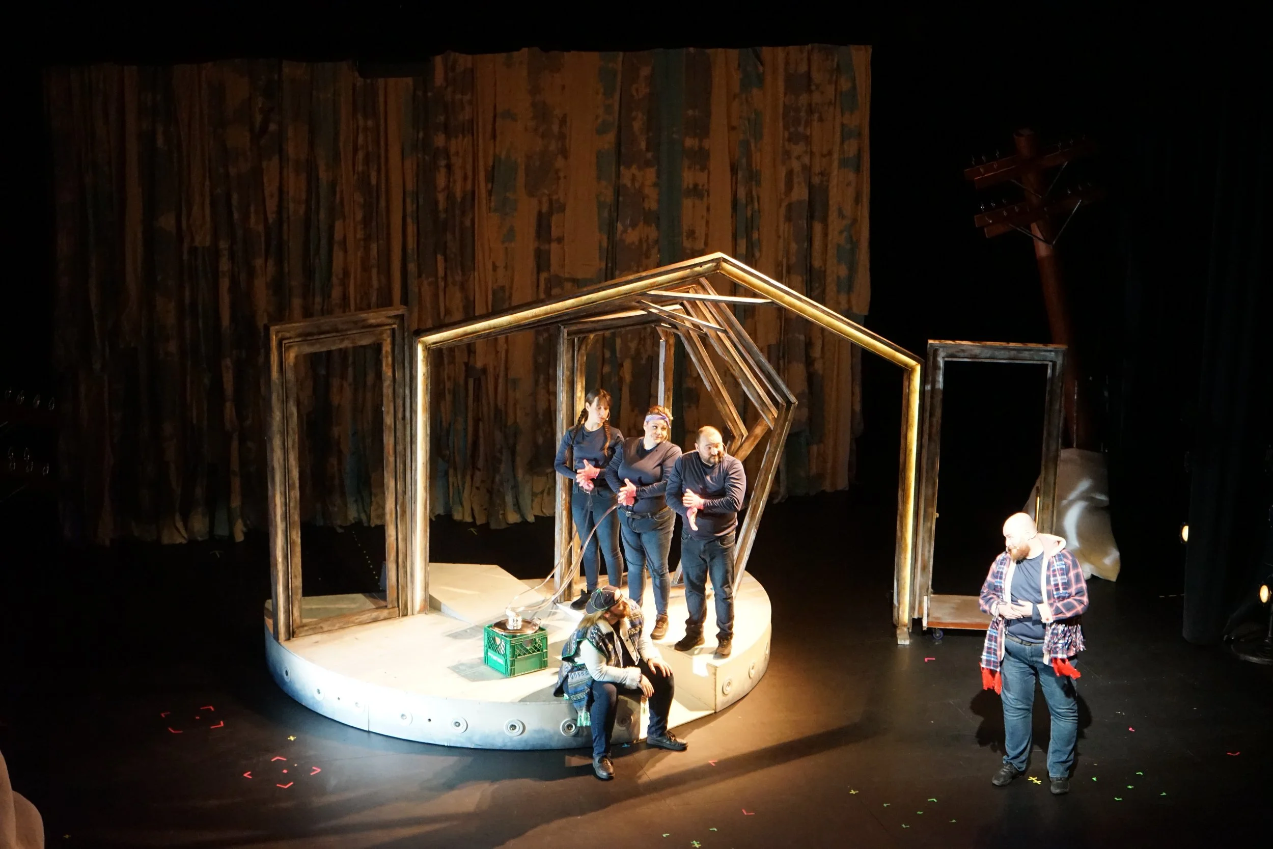 Scène de théâtre avec cinq acteurs sur une scène en bois, décor minimaliste de cadres en bois, deux acteurs assis et trois debout en train de parler, un autre acteur à droite regarde vers le bas.