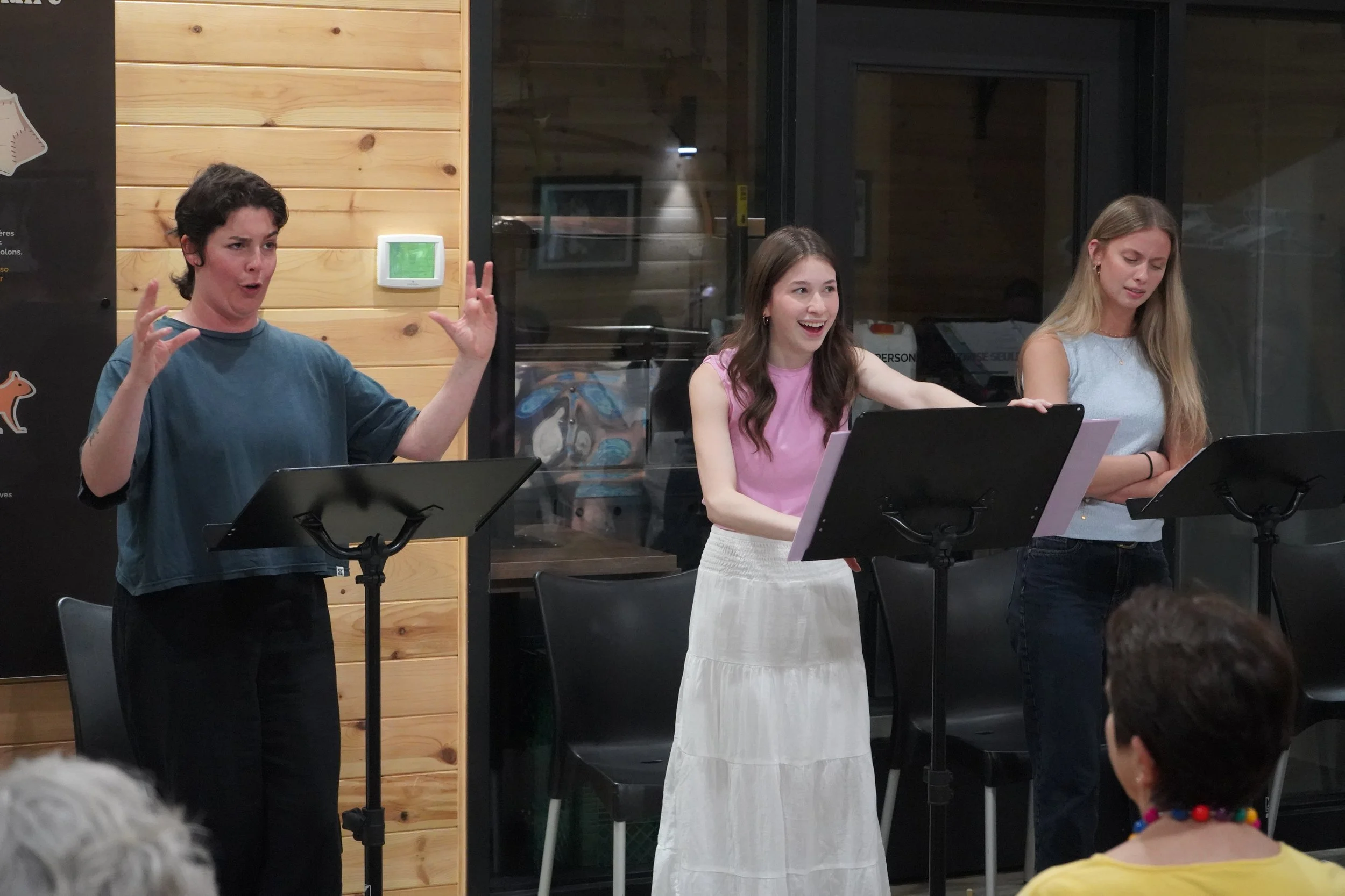 Trois femmes sont en train de faire une performance ou un discours devant un microphone. La femme à gauche gesticule avec les mains, celle au centre rit, et celle à droite lit ou chante. Elles sont dans une salle avec un décor en bois derrière eux.