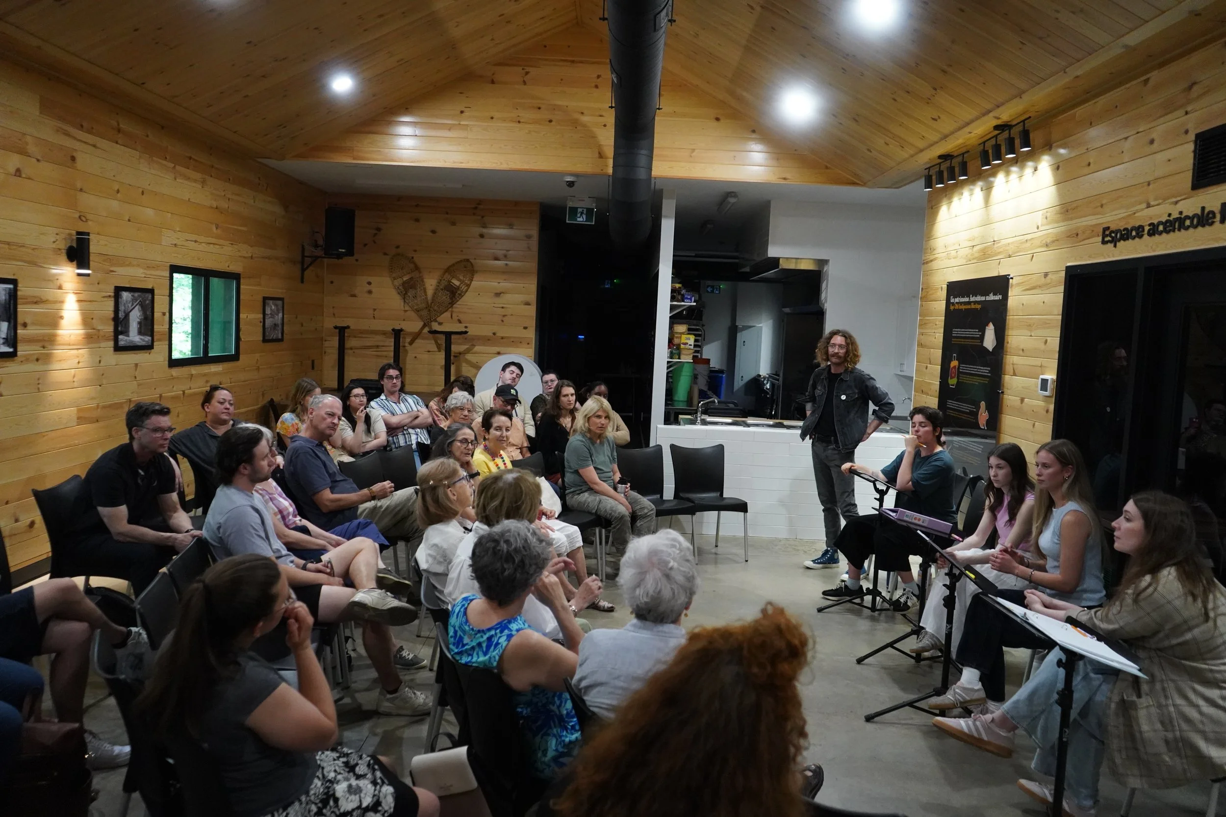 Une salle de réunion en bois avec des personnes assises et un homme en costume debout devant elles, en train de parler.