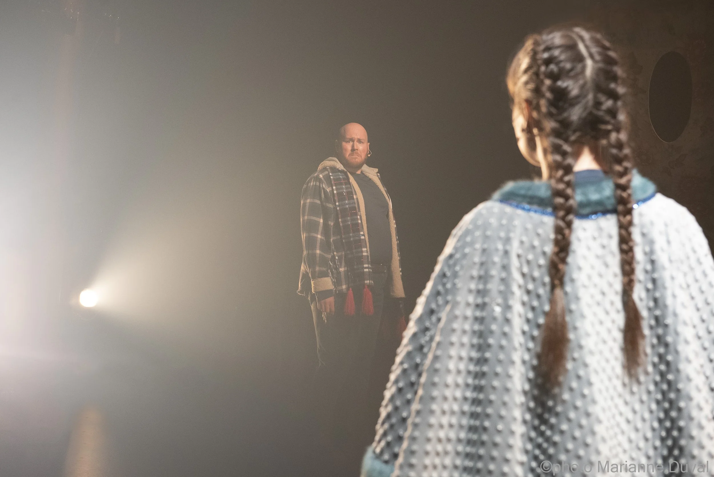 Deux personnes, un homme et une femme, se font face dans un espace sombre, probablement une scène de théâtre ou de spectacle, éclairés par une lumière douce.