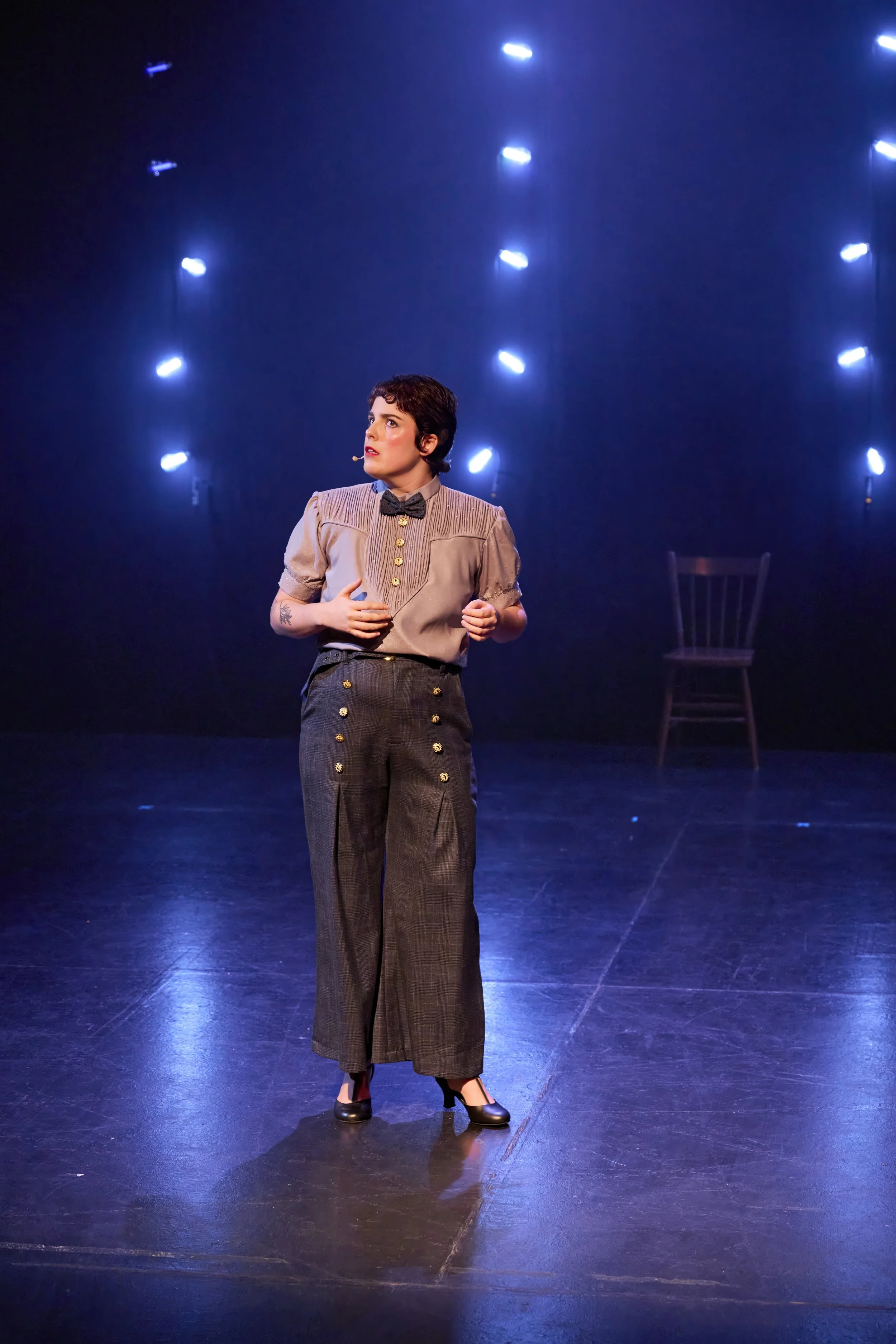 Créations In Vivo. Une femme en costume vintage sur scène avec un fond de lumières bleues et une chaise en arrière-plan.