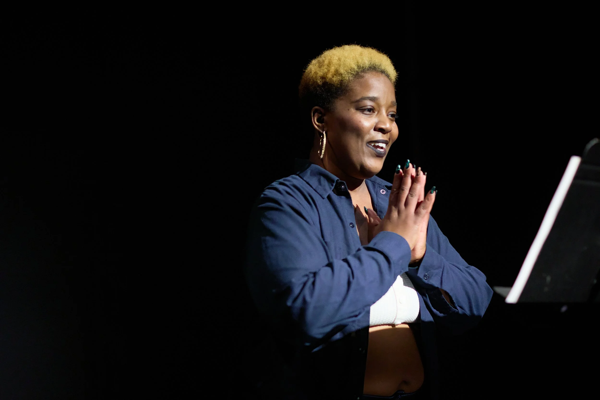 Une femme noire avec des cheveux courts blonds, en train de jouer du piano sur scène, avec ses mains jointes en position de prière, sous un éclairage focussé sur elle contre un fond noir.