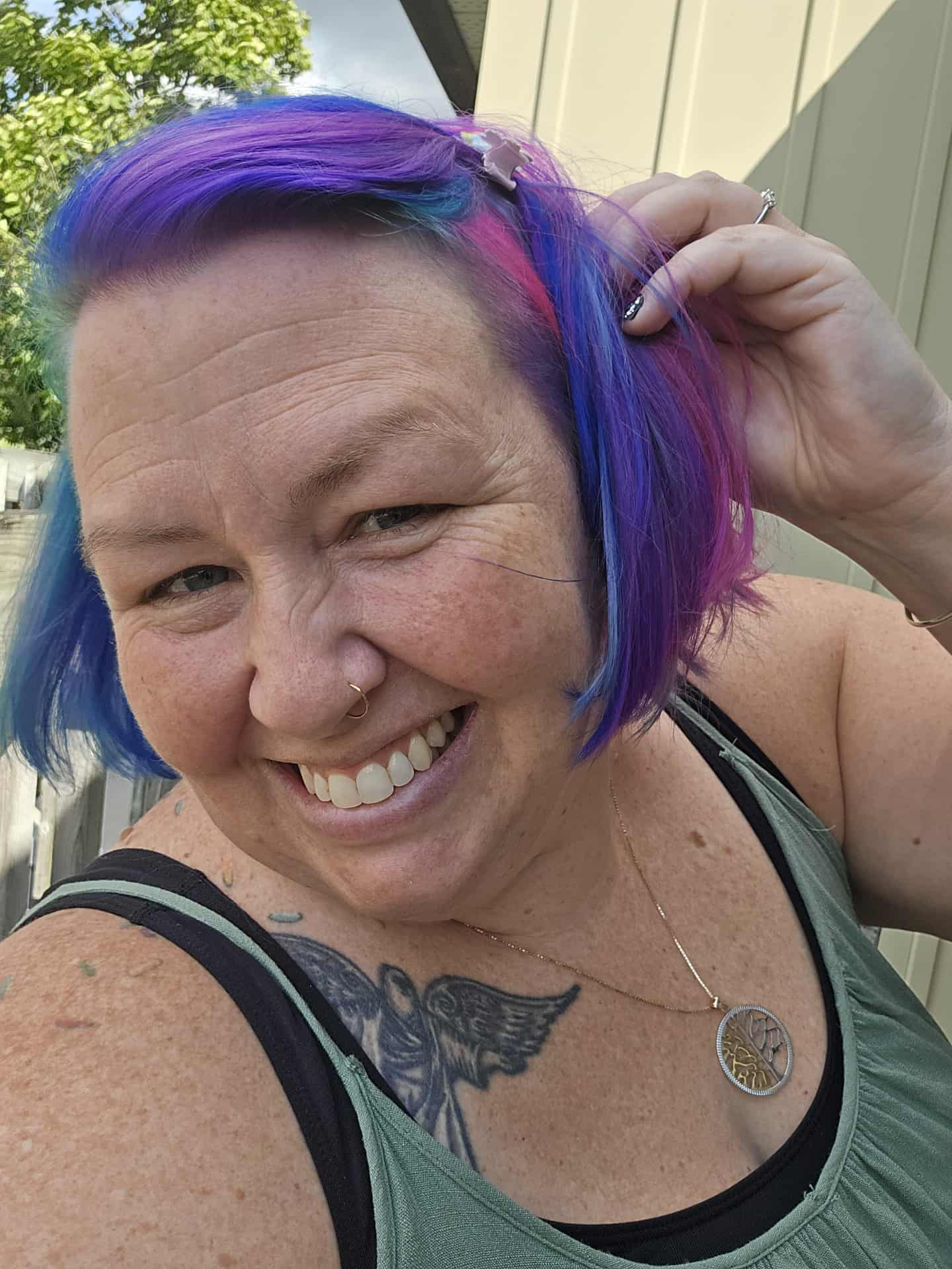 Une femme souriante avec des cheveux colorés en violet, bleu, et rose, portant un collier avec un pendentif," tatouage d'un ange sur l'épaule, posant en extérieur près d'une maison.
