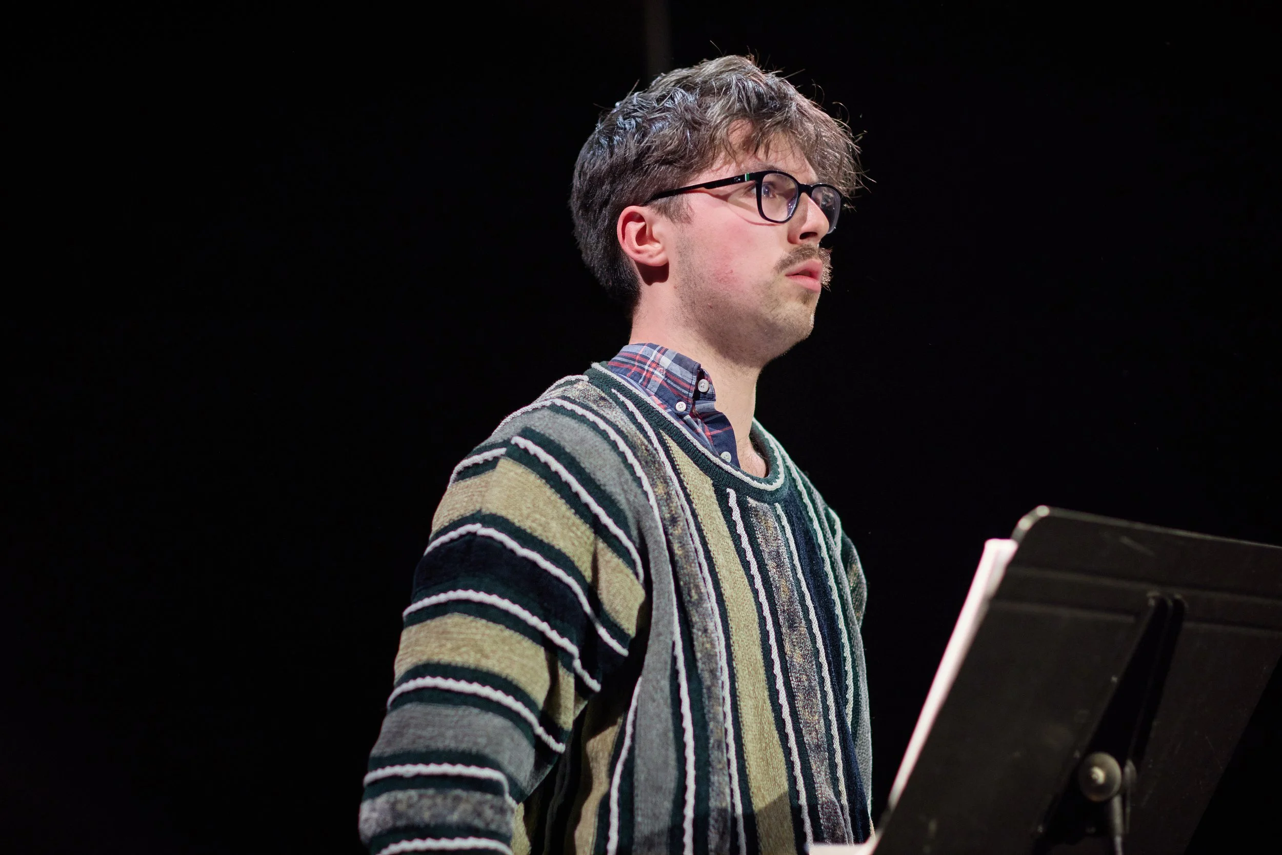 Un jeune homme portant des lunettes, une chemise à carreaux et un pull rayé, en train de lire une partition musicale, sur fond noir.