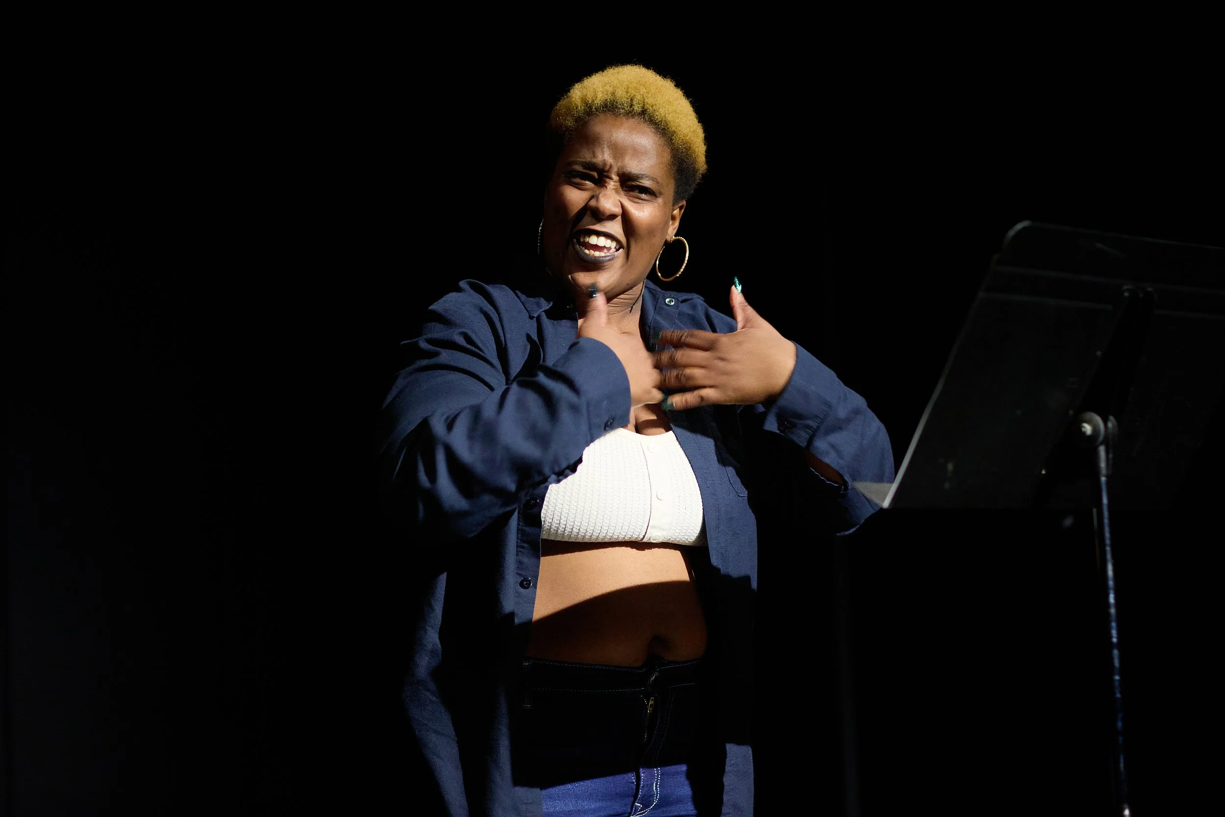 Une femme expressivement en train de chanter ou de parler, avec une main sur la gorge et une autre sur la poitrine, portant une chemise bleue ouverte sur un haut blanc, avec des boucles d'oreilles larges, dans un environnement sombre.