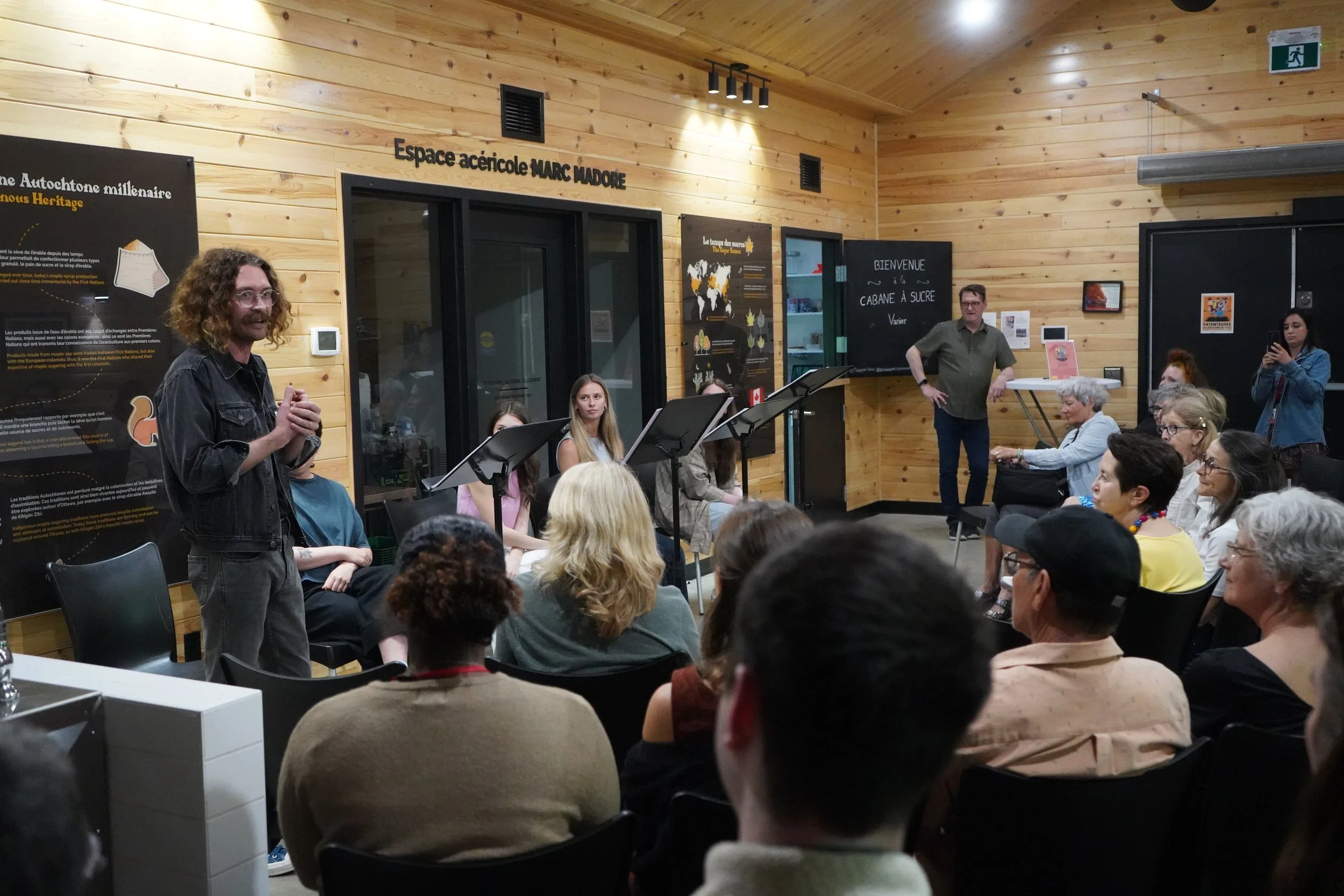 Une salle avec un groupe de personnes assises, un homme à gauche parle et d'autres personnes sont au fond devant un tableau noir avec du texte et une lecture sur des chèvres ou des animaux. La salle a des murs en bois et une ambiance chaleureuse.