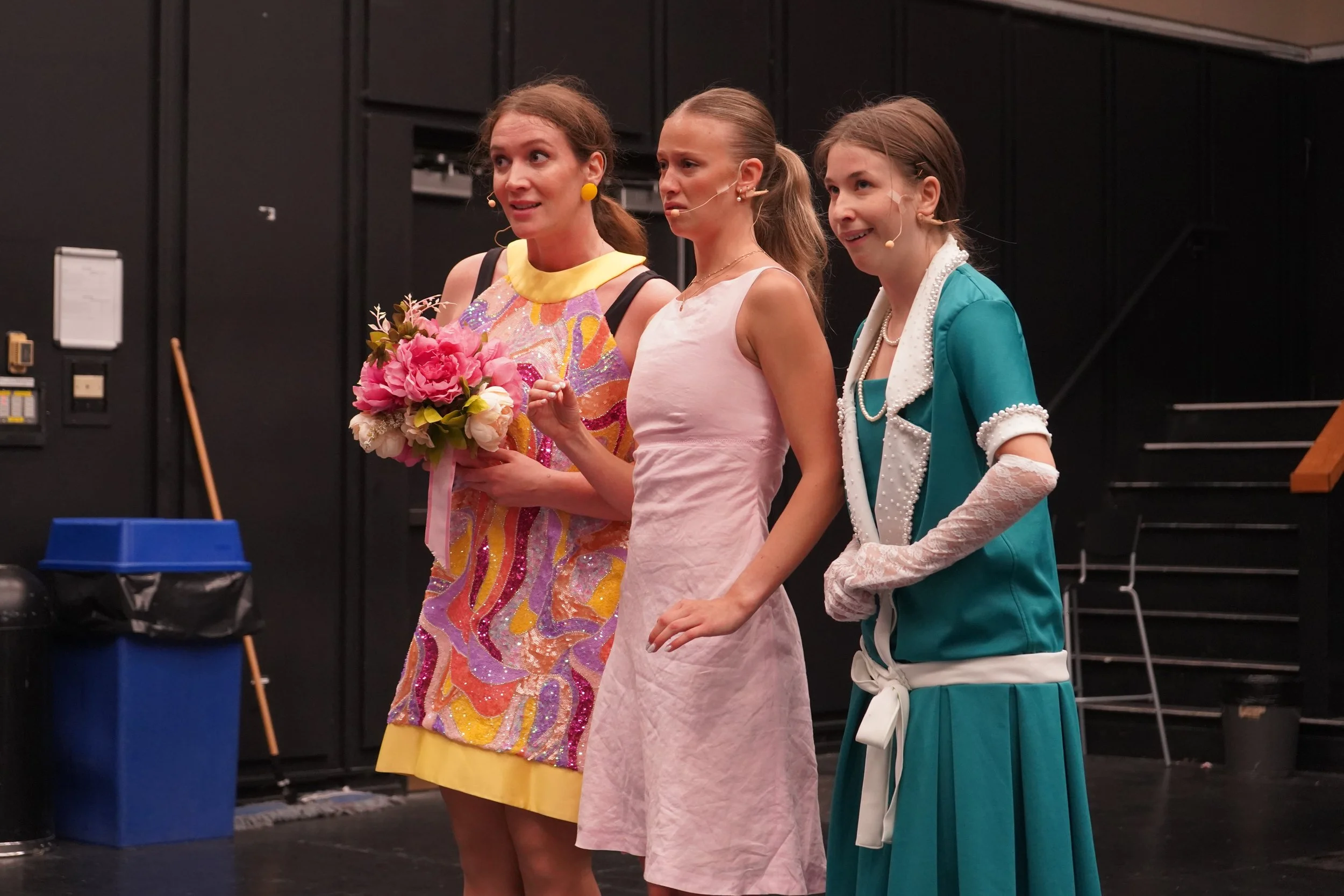 Trois femmes en scène, la première tient un bouquet de fleurs et porte une robe colorée, la deuxième porte une robe rose pâle, et la troisième une veste turquoise avec une jupe blanche, toutes avec des micros cousus sur leurs visages, dans un décor d