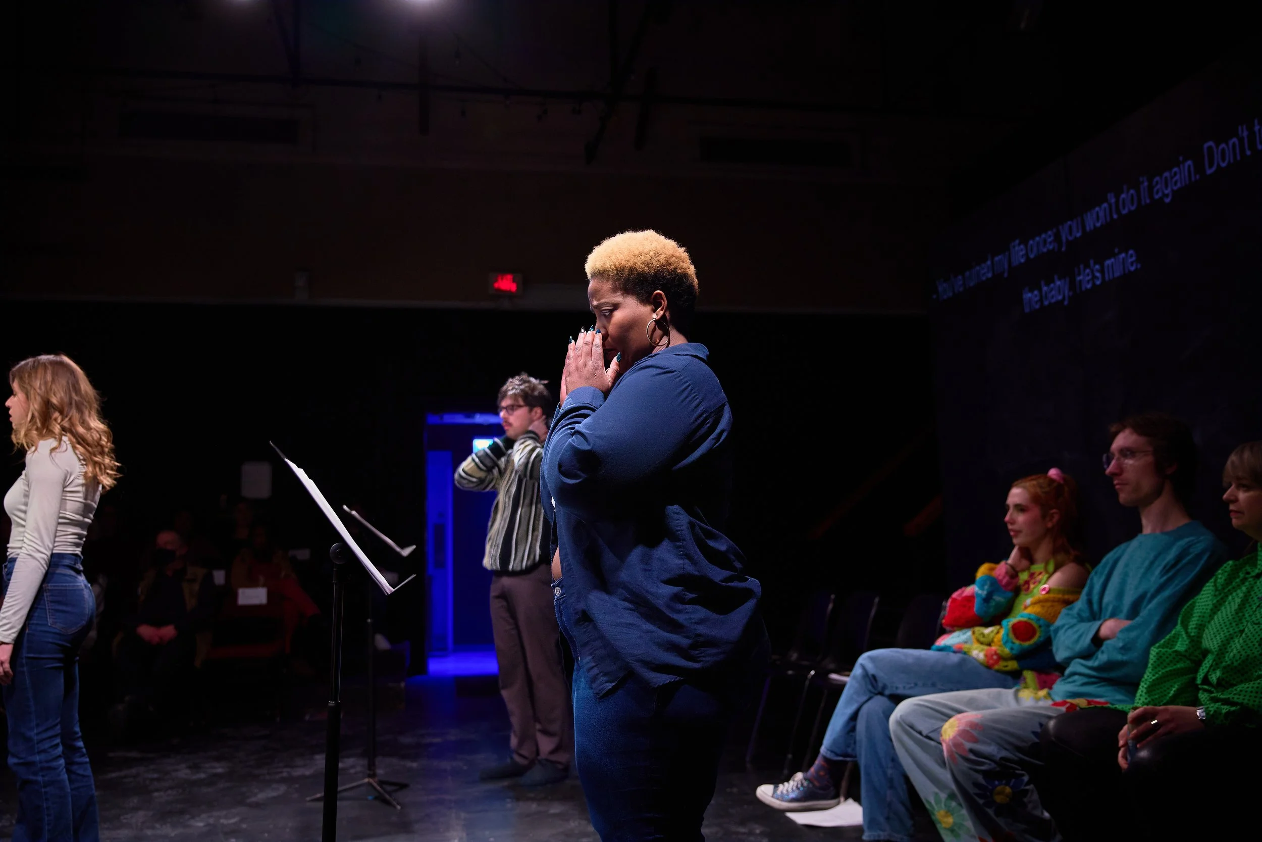 Un groupe de personnes en théâtre, une femme en bleu avec une coupe de cheveux courte et blonde est en prière ou méditation, entourée d'autres personnes assises ou debout, dans une ambiance sombre avec un écran en arrière-plan affichant des sous-titr