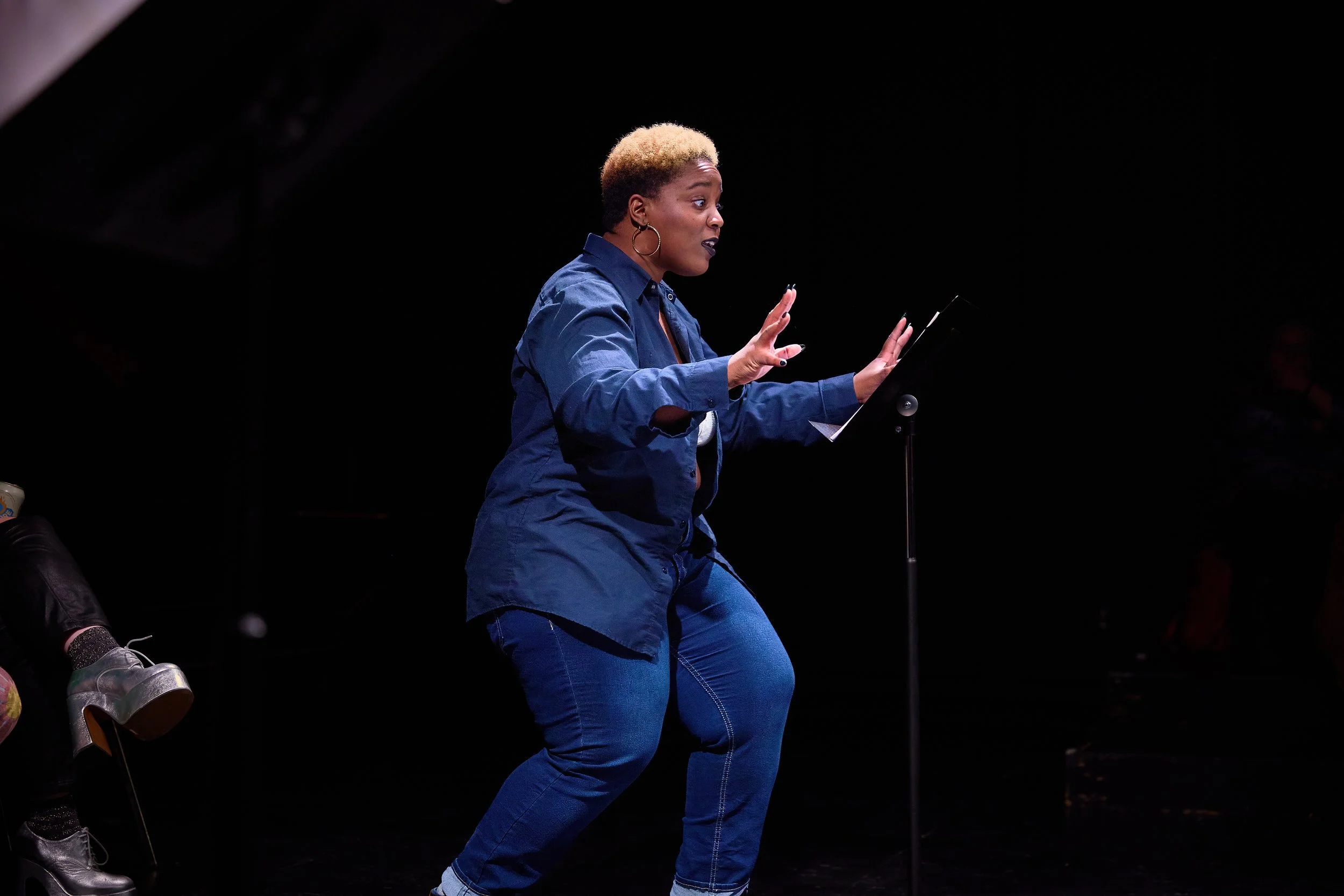 Une femme en jean et chemise bleue se tient devant un pupitre, avec une expression de surprise ou d'inquiétude, dans un environnement sombre. 