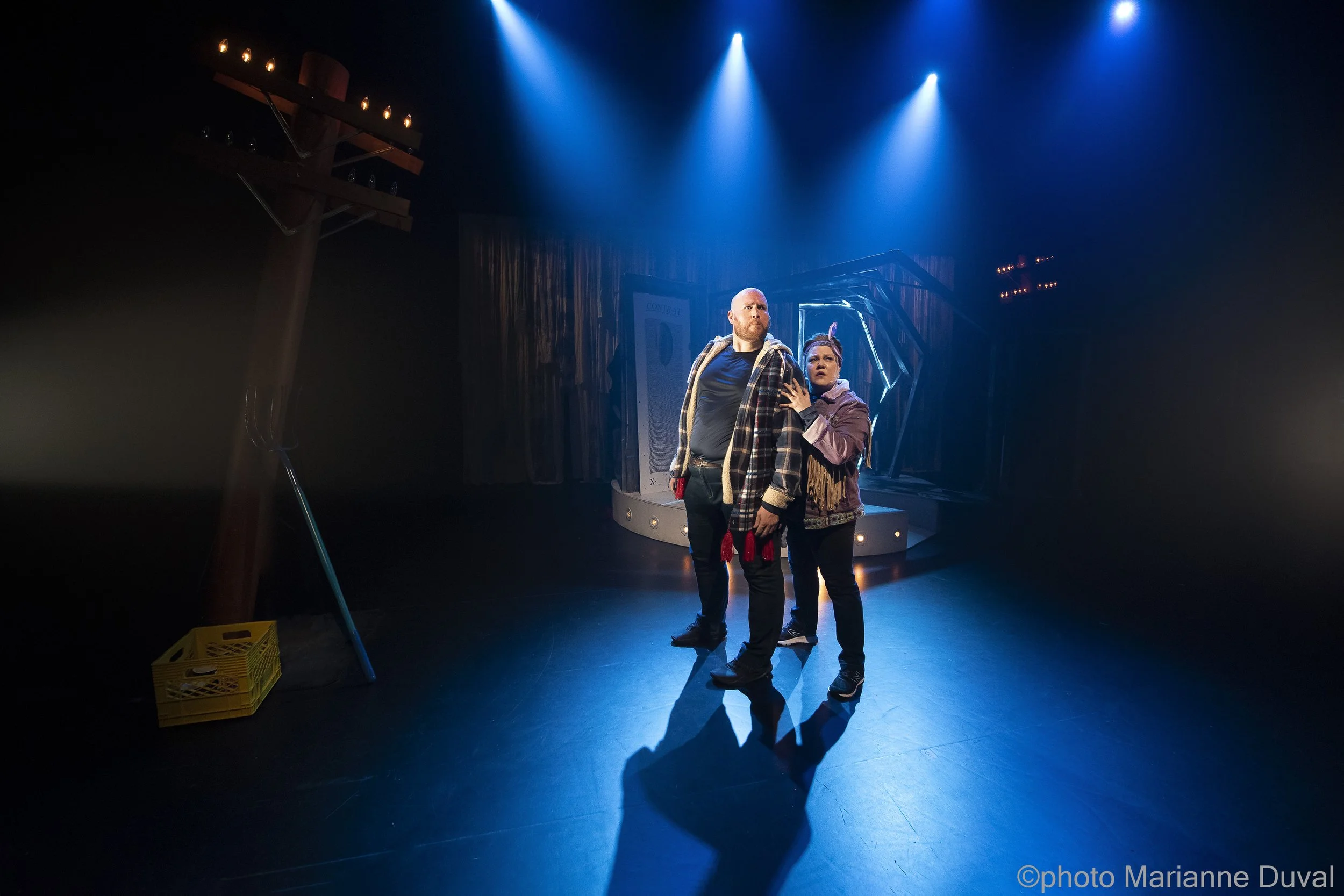 Deux acteurs sur scène dans une pièce de théâtre, éclairés par des projecteurs bleus, avec un décor minimaliste en arrière-plan.