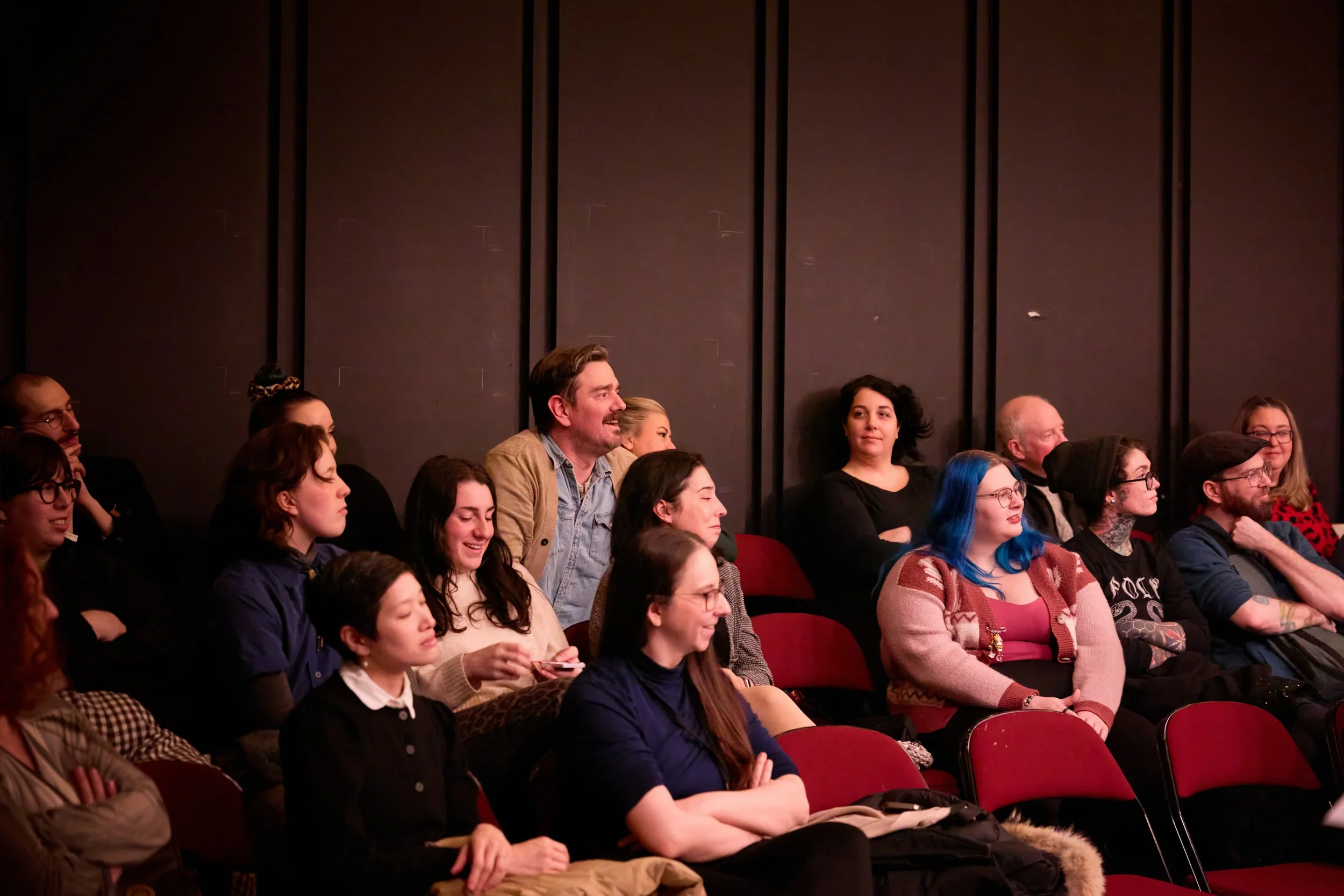 Groupe de personnes assises dans une salle de spectacle ou de théâtre, regardant vers la scène avec des expressions diverses. Certains sourient, d'autres semblent attentifs ou concentrés.
