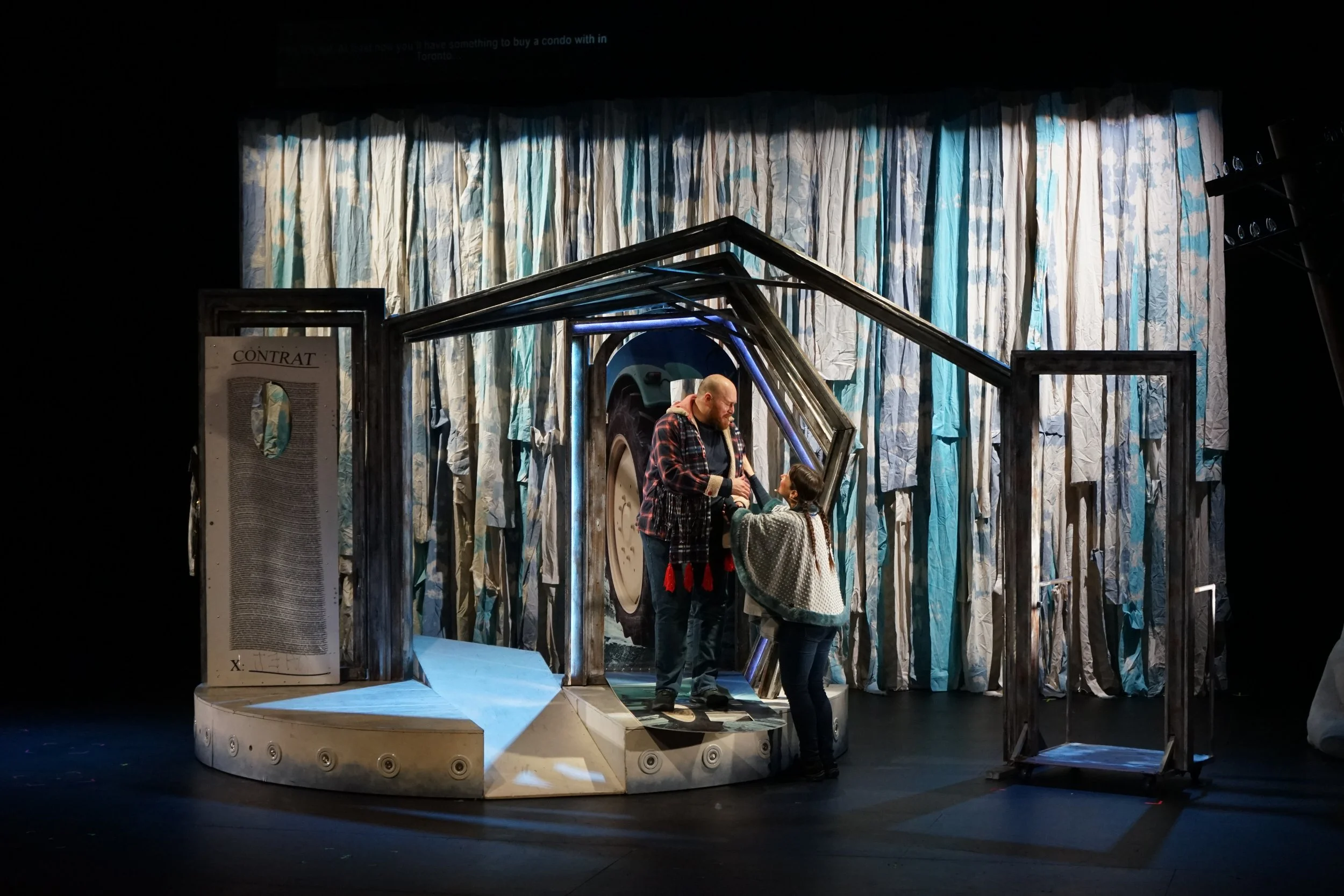 Scène théâtrale avec deux acteurs, un homme et une femme, dans une mise en scène représentant une maison avec un mur en papier froissé et une grande roue derrière une structure métallique.