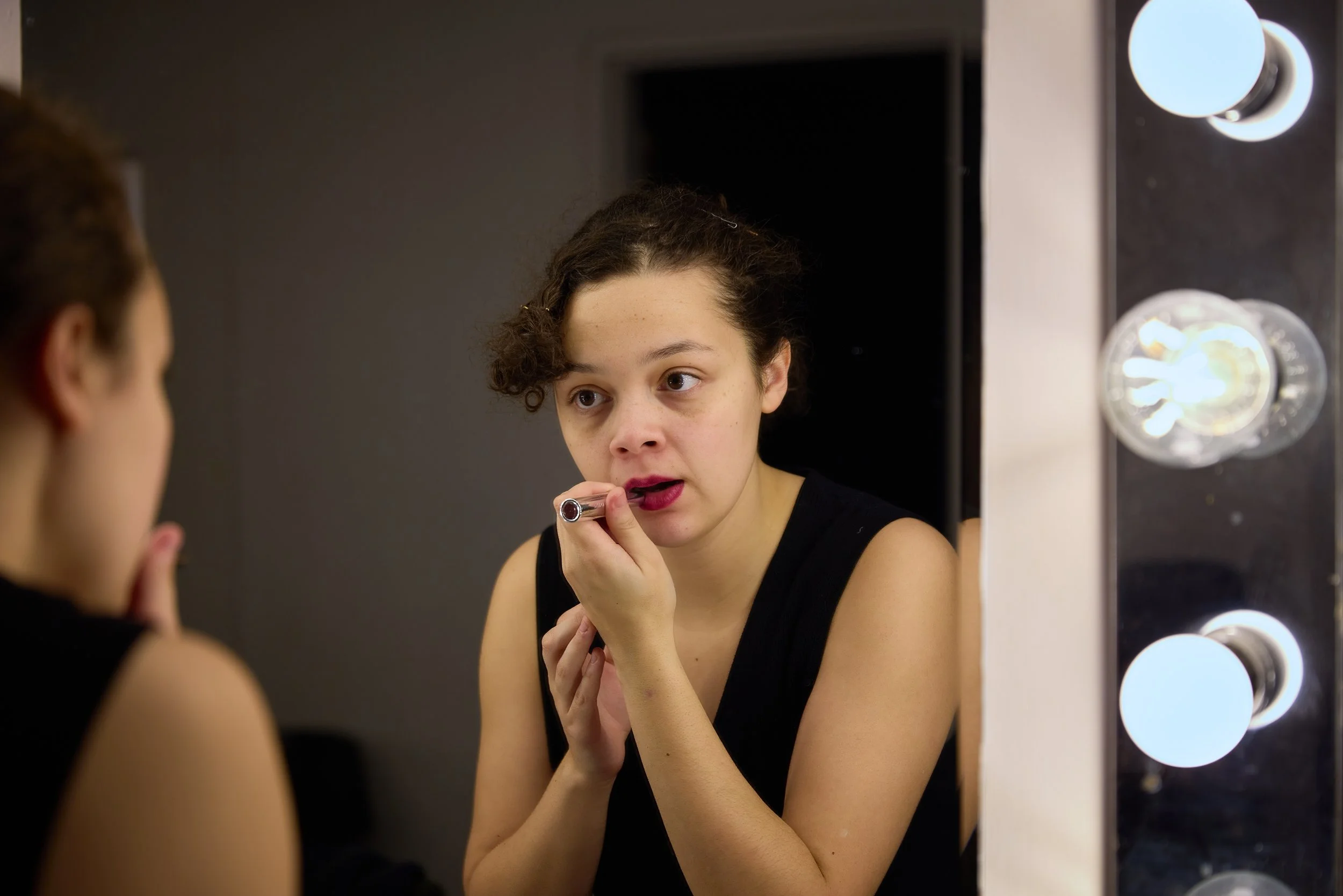 Une jeune femme se maquille devant un miroir éclairé par des ampoules, avec le reflet visible sur la vitre.