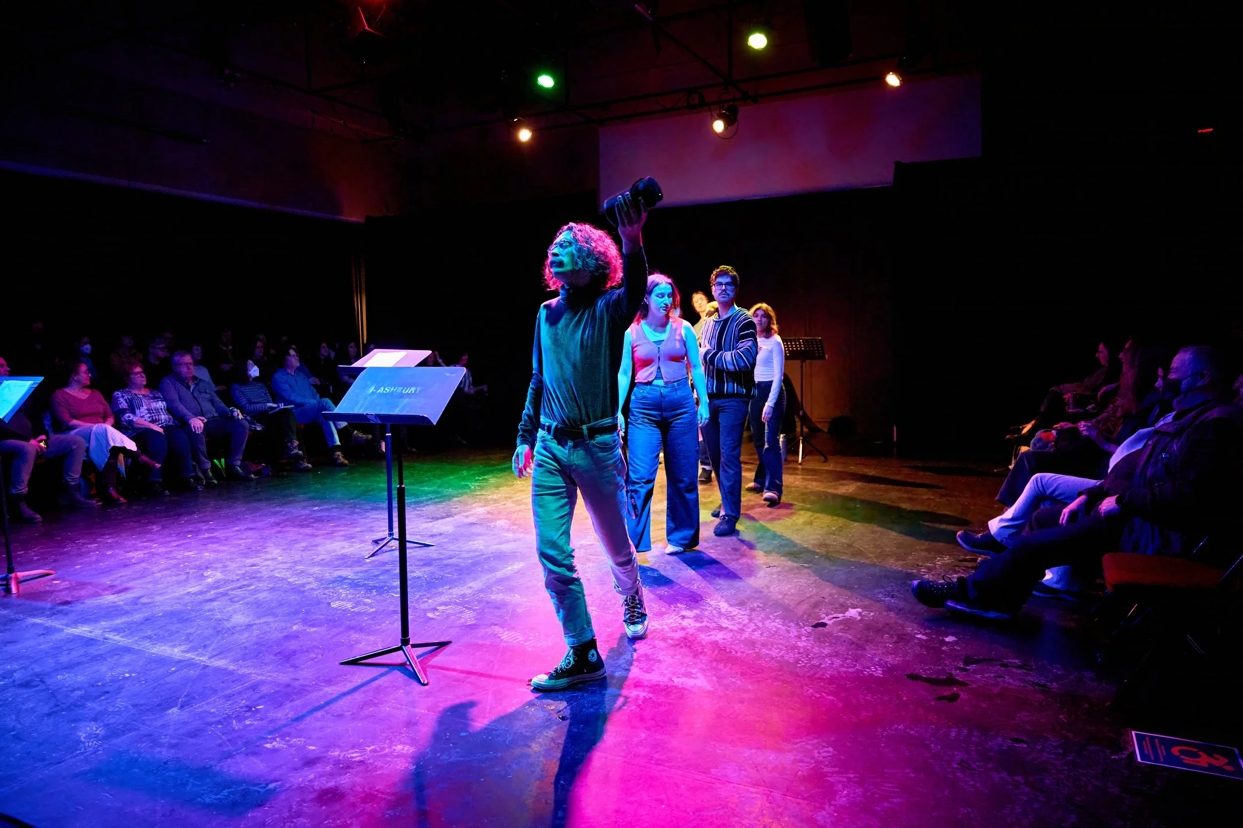 Actrice dans une pièce de théâtre ou une performance, debout sur scène sous des lumières colorées, devant un micro, avec plusieurs personnes en ligne devant un public dans une salle sombre.