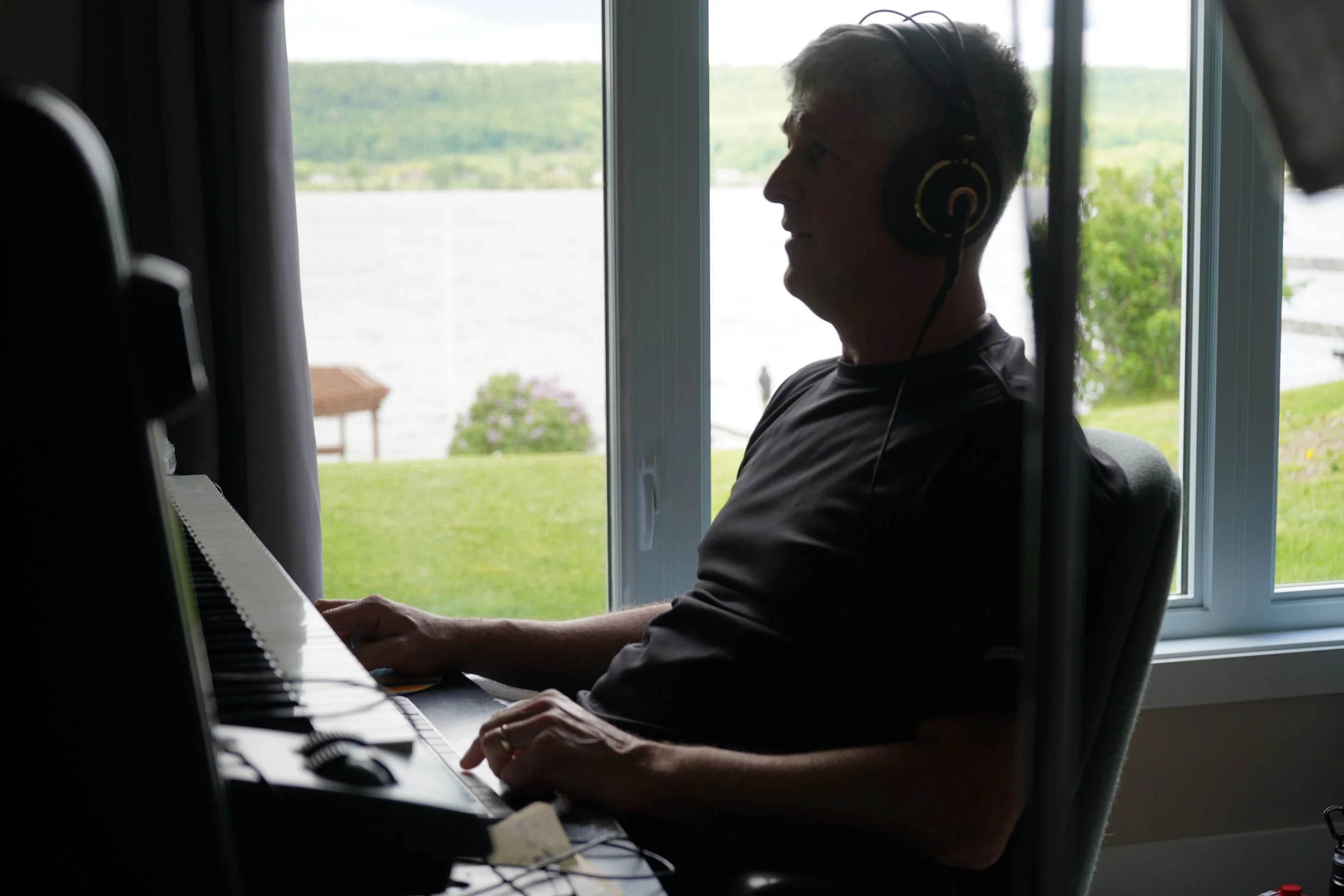 Un homme âgé joue du piano dans une pièce près d'une grande fenêtre avec vue sur un lac et des arbres.
