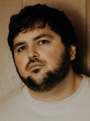 Jeune homme avec une barbe, regardant vers l'objectif, portant un T-shirt blanc, fond en bois.
