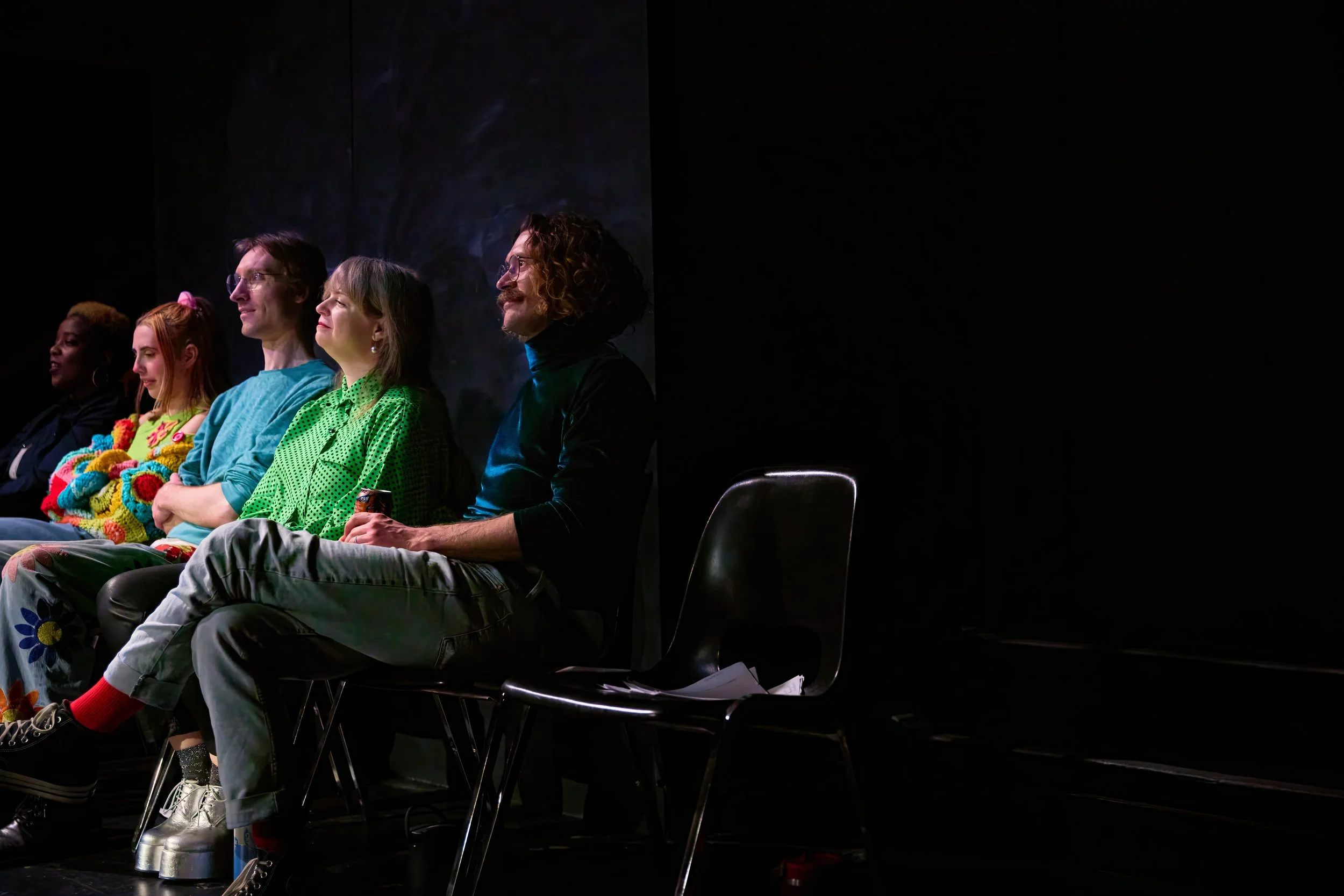Groupe de personnes assises en ligne, dans un endroit sombre, probablement un théâtre ou une salle de spectacle.