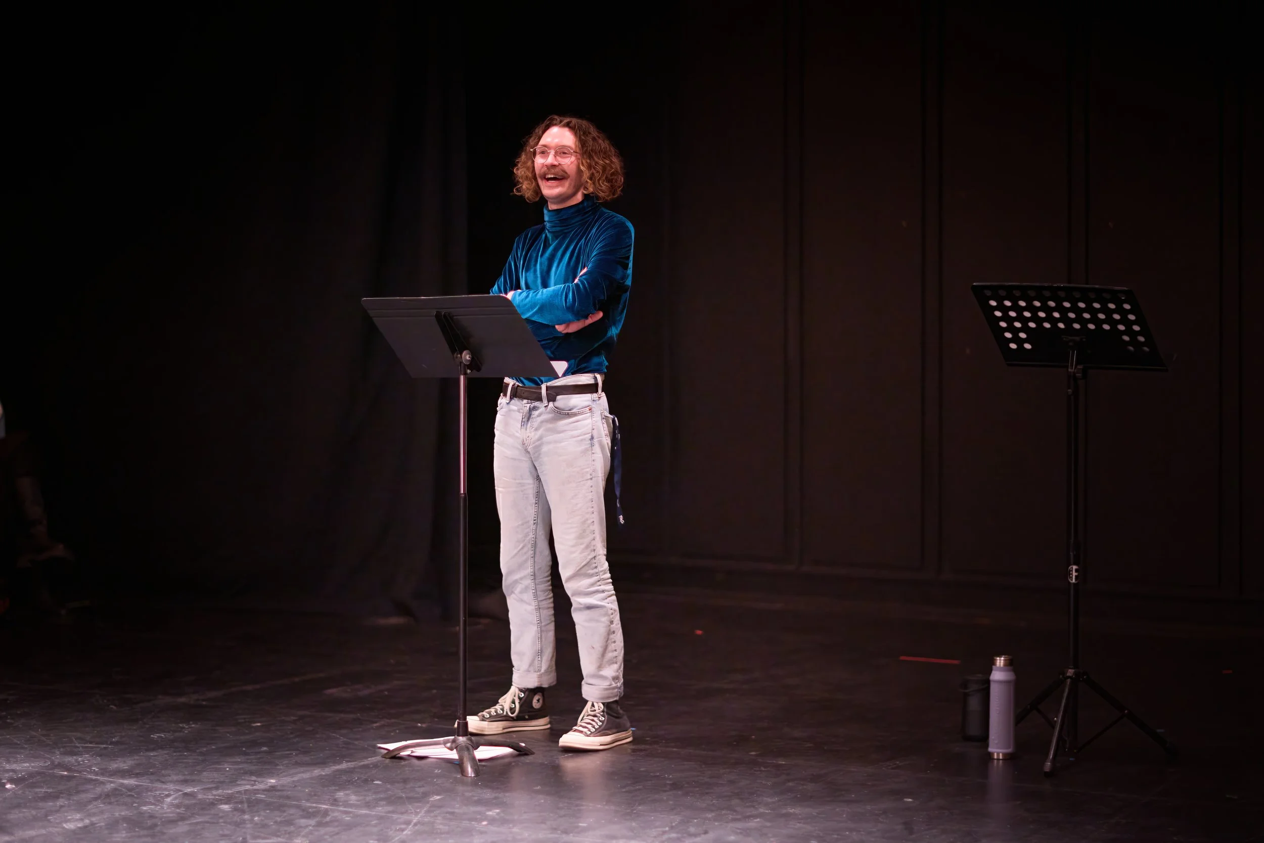 Homme avec des cheveux bouclés et une moustache, portant un pull en velours bleu et un jean clair, riant et avec les bras croisés, se tenant devant un pupitre sur scène, avec des chevalets pour la lecture de partition ou texte situés à côté.