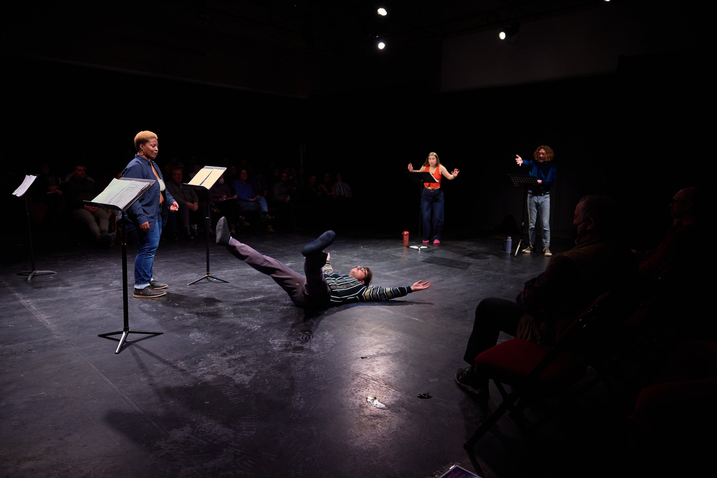 Une scène de théâtre ou de spectacle avec un homme au sol faisant un mouvement de danse ou de théâtre, et trois femmes debout, probablement en train de performer ou de donner des indications, avec un public en arrière-plan, dans une salle sombre avec