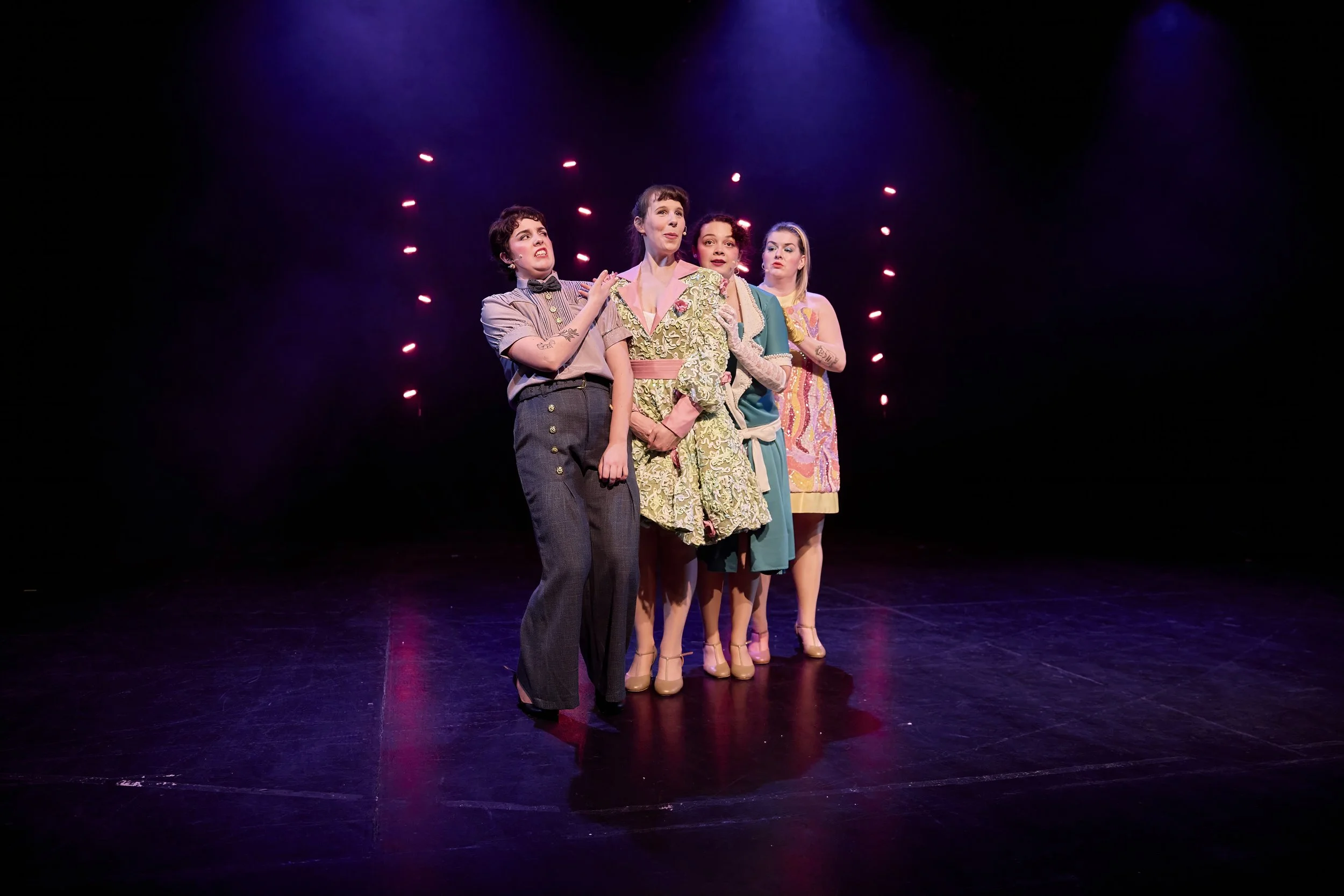 Créations In Vivo. Quatre femmes en costume se tenant sur scène sous des lumières violettes.