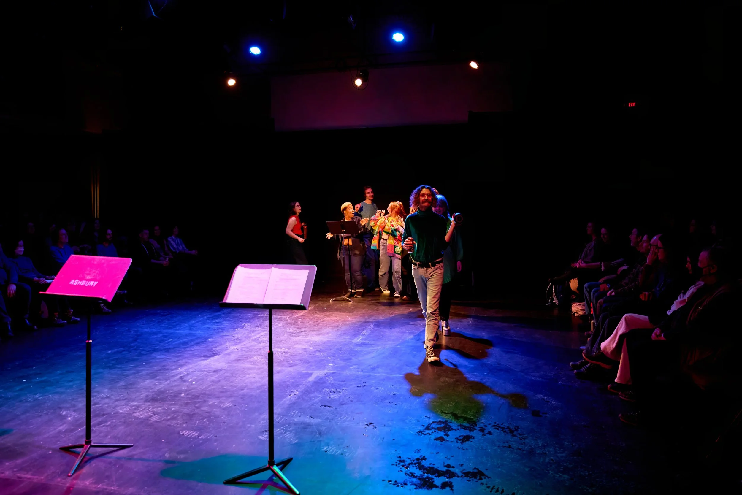 Scène de théâtre avec plusieurs acteurs sur scène, un public assis sur les côtés, musique et ambiance de spectacle.