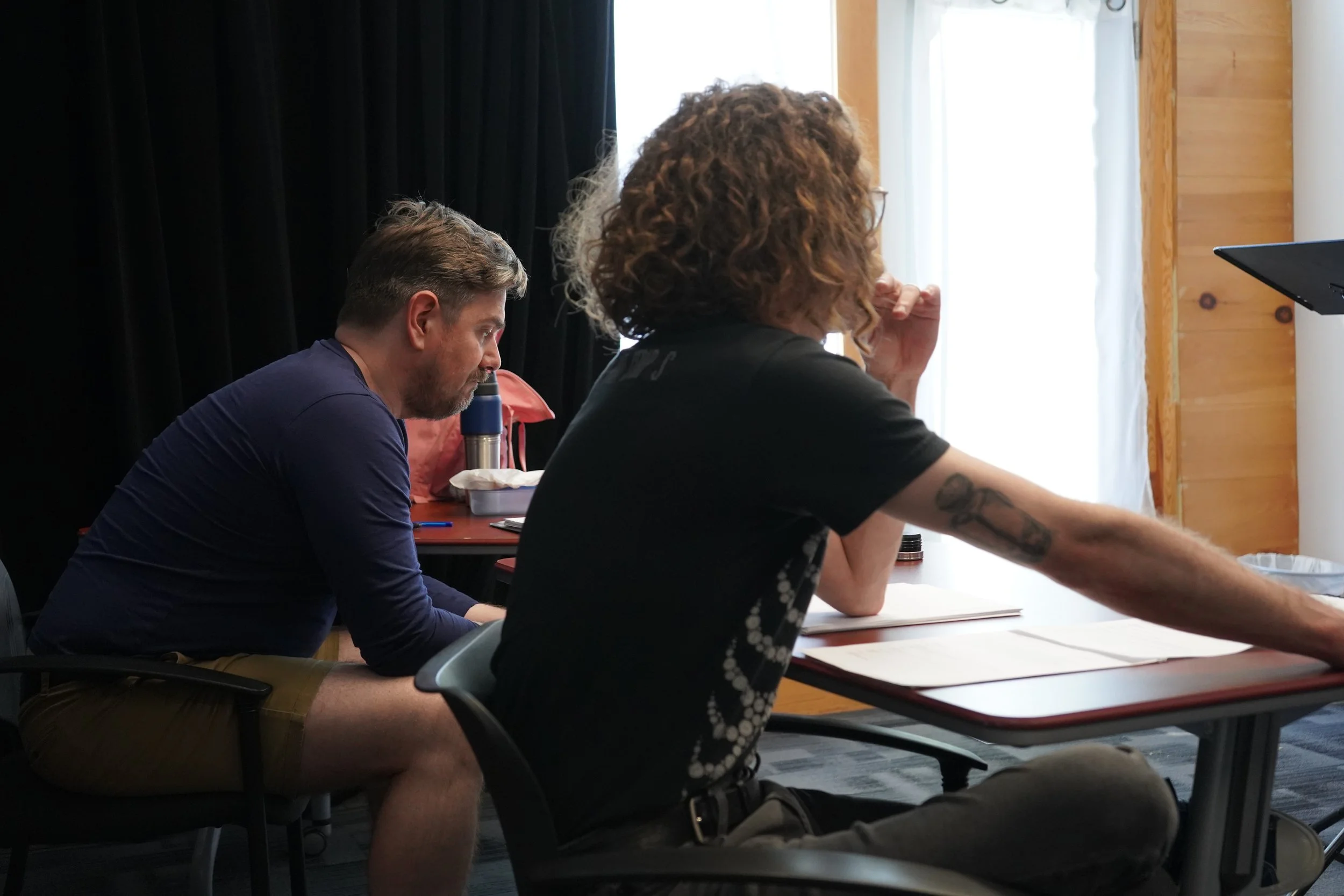 Deux hommes assis à une table lors d'une réunion ou d'un atelier, l'un avec un micro et l'autre regardant un écran, dans une pièce avec fenêtre et rideaux blancs.