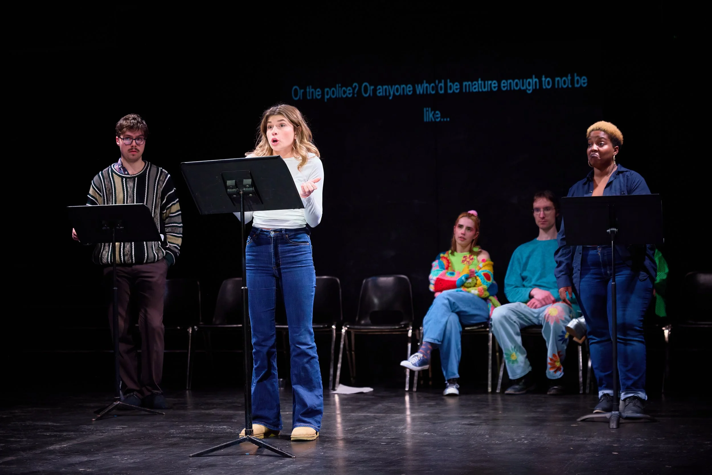 Groupe de cinq jeunes sur scène, l'un parlant avec un micro, fond noir avec sous-titres en anglais.