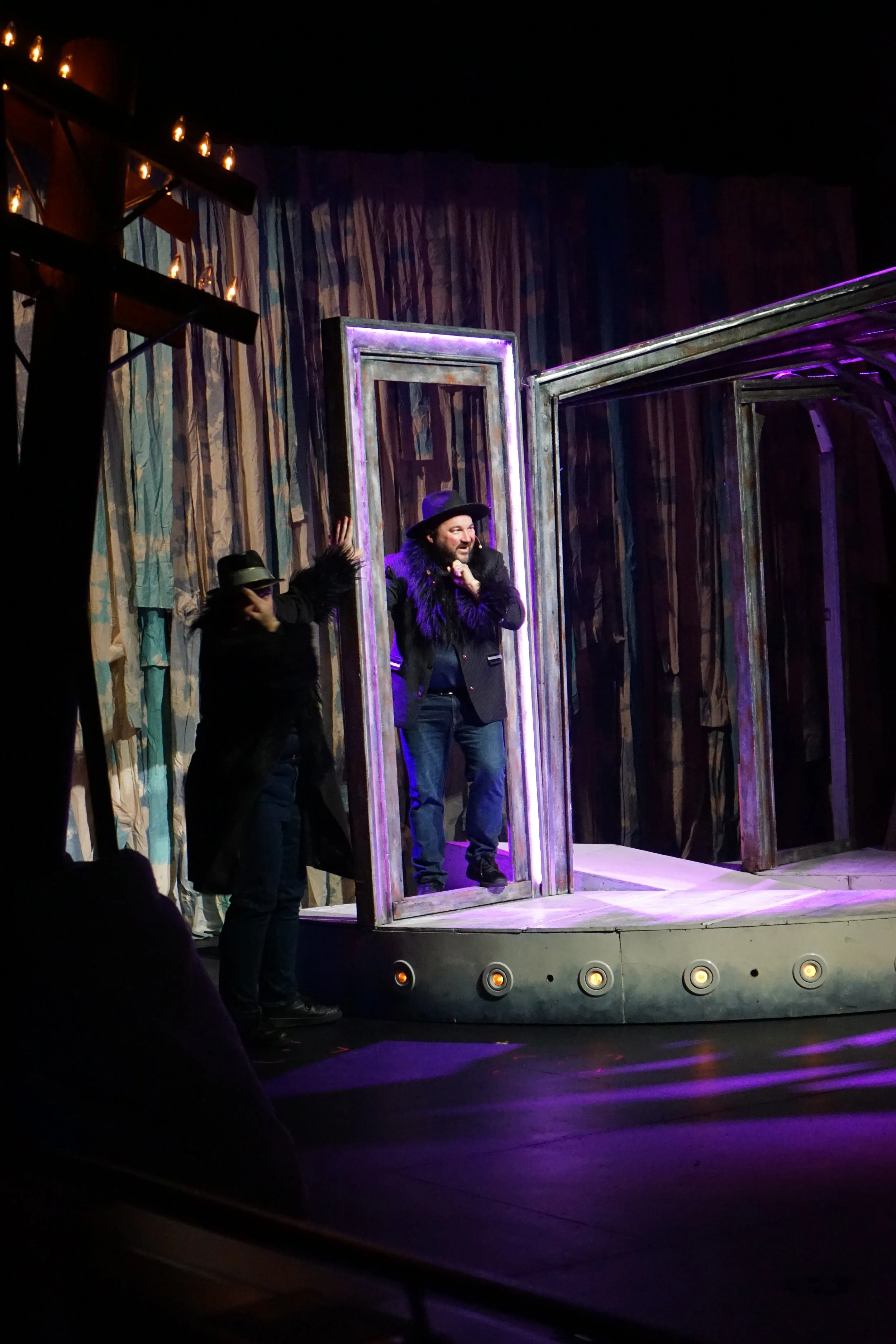 Une scène de théâtre avec un homme portant un chapeau et un manteau à col en fourrure à l'intérieur d'un cadre en bois, tandis qu'une autre personne se tient à côté, dans un décor avec des rideaux colorés.