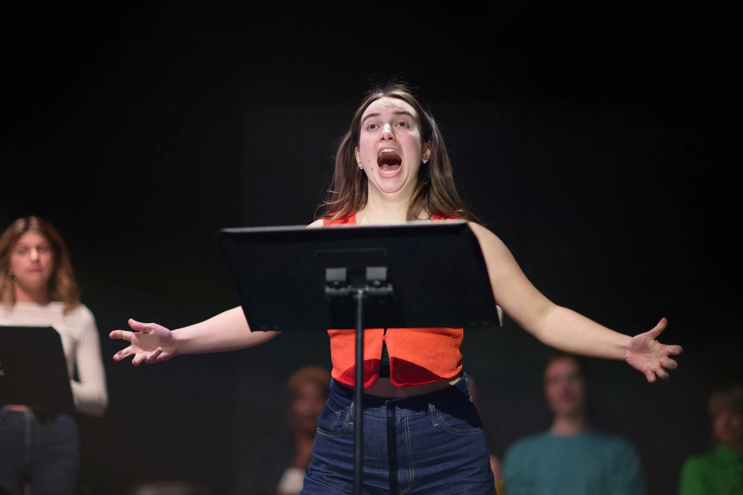Une femme chante lors d'une performance ou d'une répétition, avec d'autres personnes en arrière-plan, dans un espace sombre.
