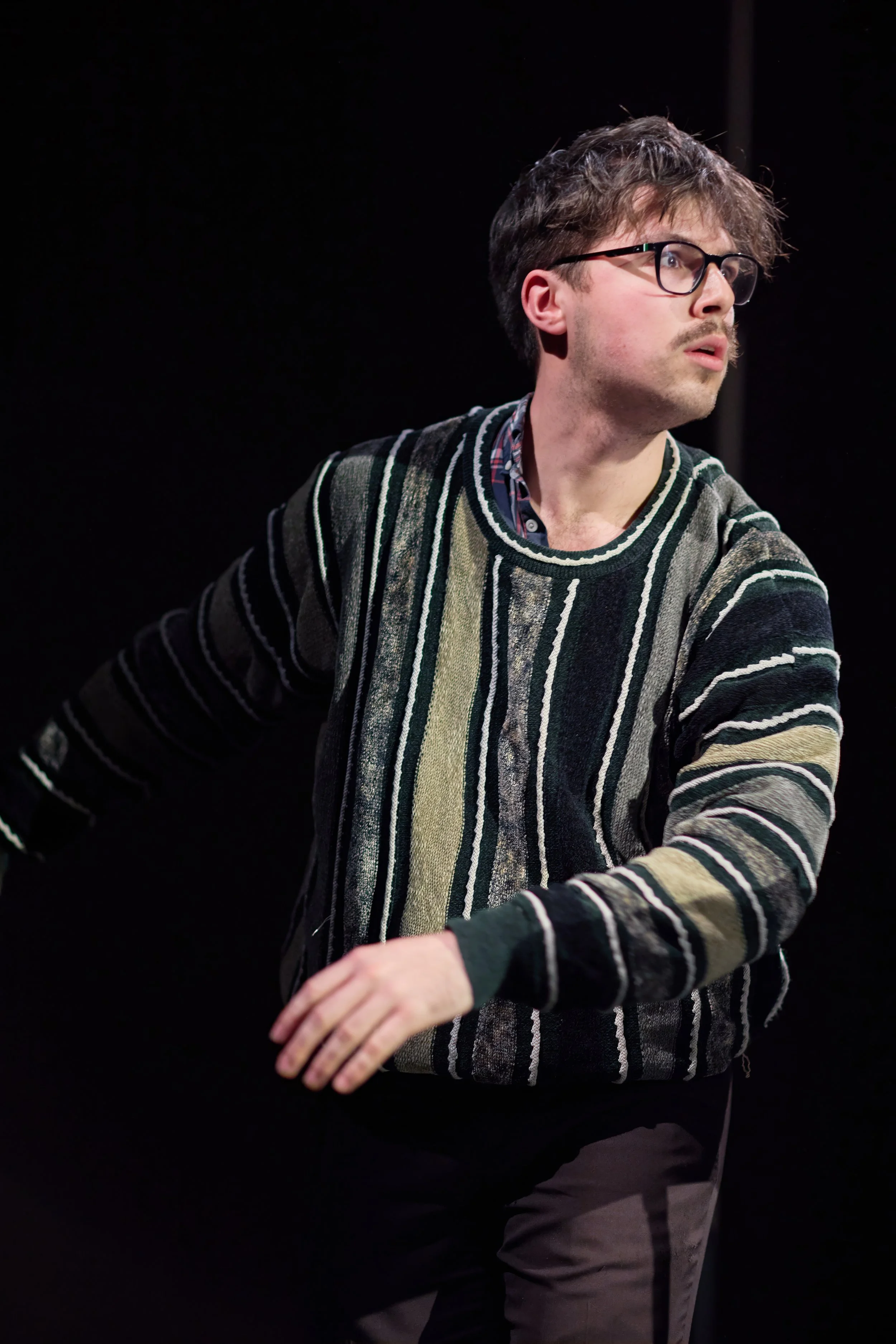 Un homme avec des lunettes, portant un pull à rayures, semble être en train de jouer la comédie sur scène, avec un fond noir.