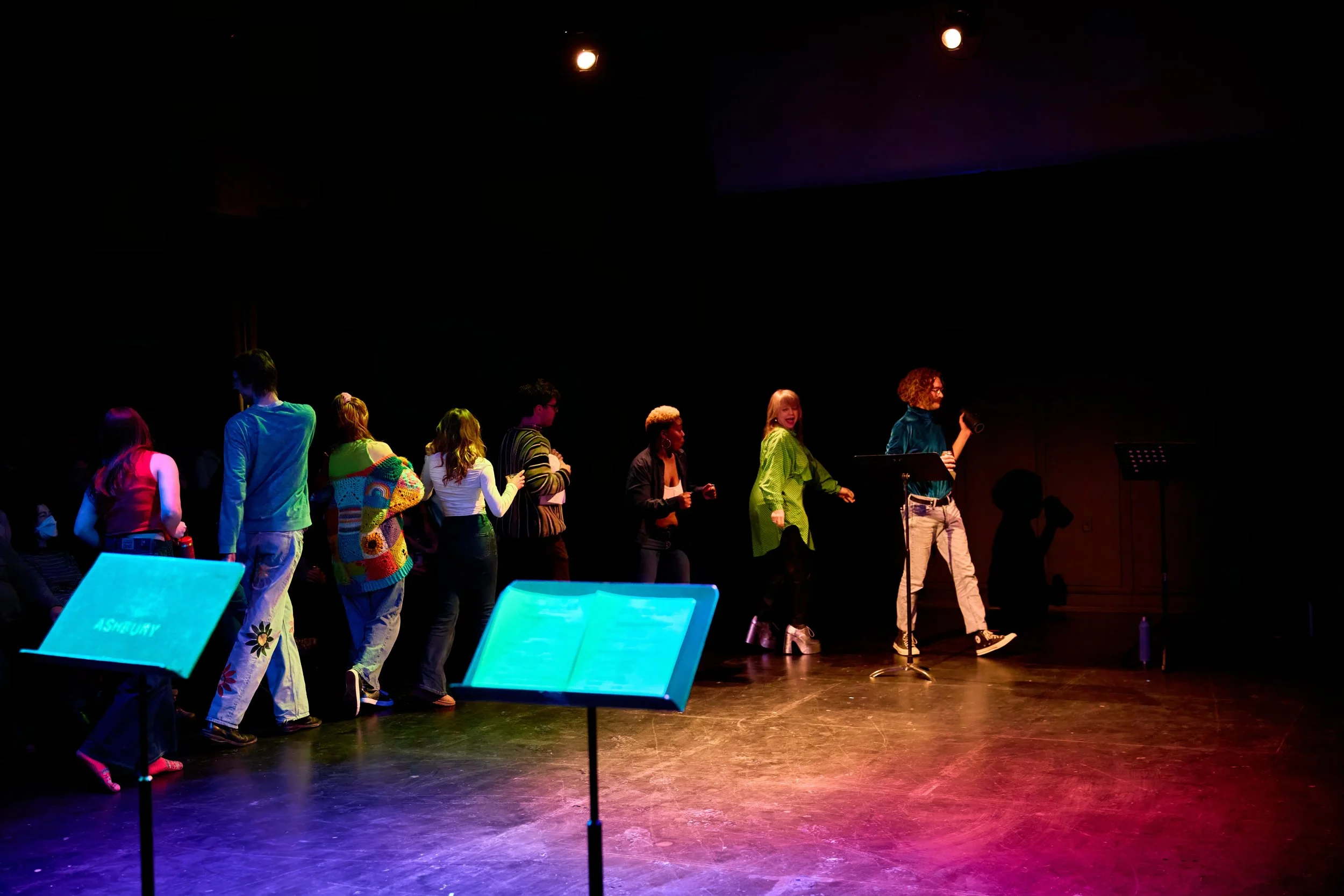 Groupe de personnes se tenant en file sur une scène sombre, avec un musicien au micro et des pupitres lumineux en premier plan, dans une atmosphère de spectacle ou de répétition.