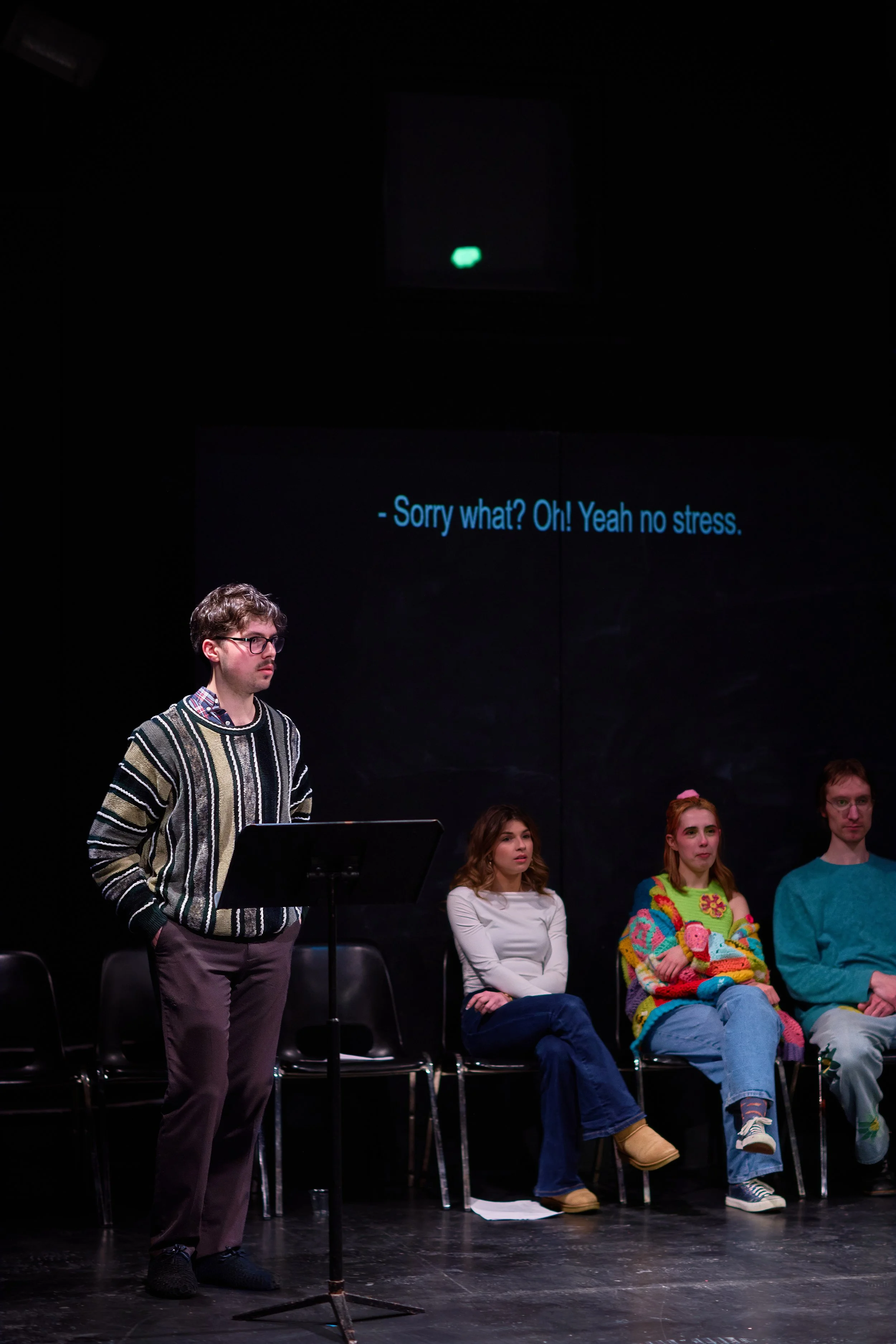Un groupe de personnes assises sur des chaises sur scène, avec un homme debout à une lecture ou une performance, et une projection à l'arrière avec des sous-titres en anglais