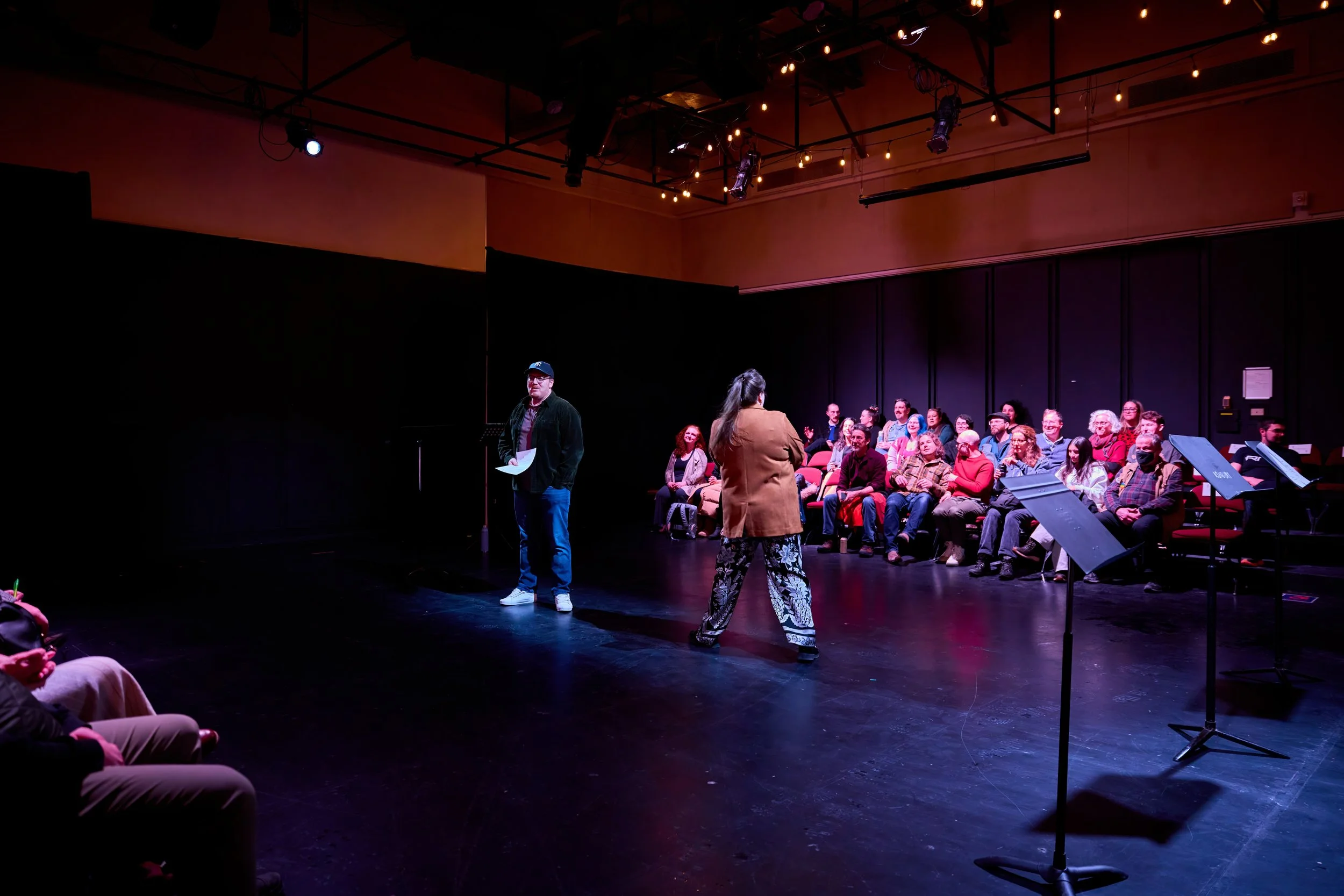 Une scène de théâtre avec deux acteurs au centre, un homme et une femme, et un public attentif assis en arrière-plan. Le décor est minimaliste avec un éclairage tamisé.