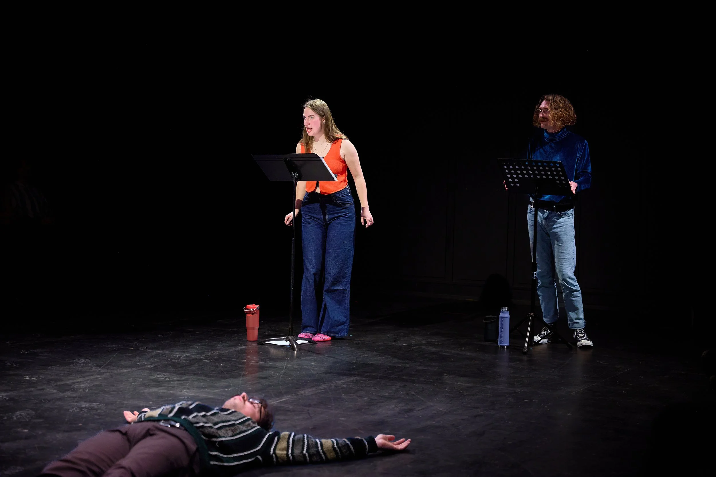 Une scène de théâtre où une femme est debout devant un micro, avec une autre femme à côté, et une personne allongée sur le sol au premier plan.