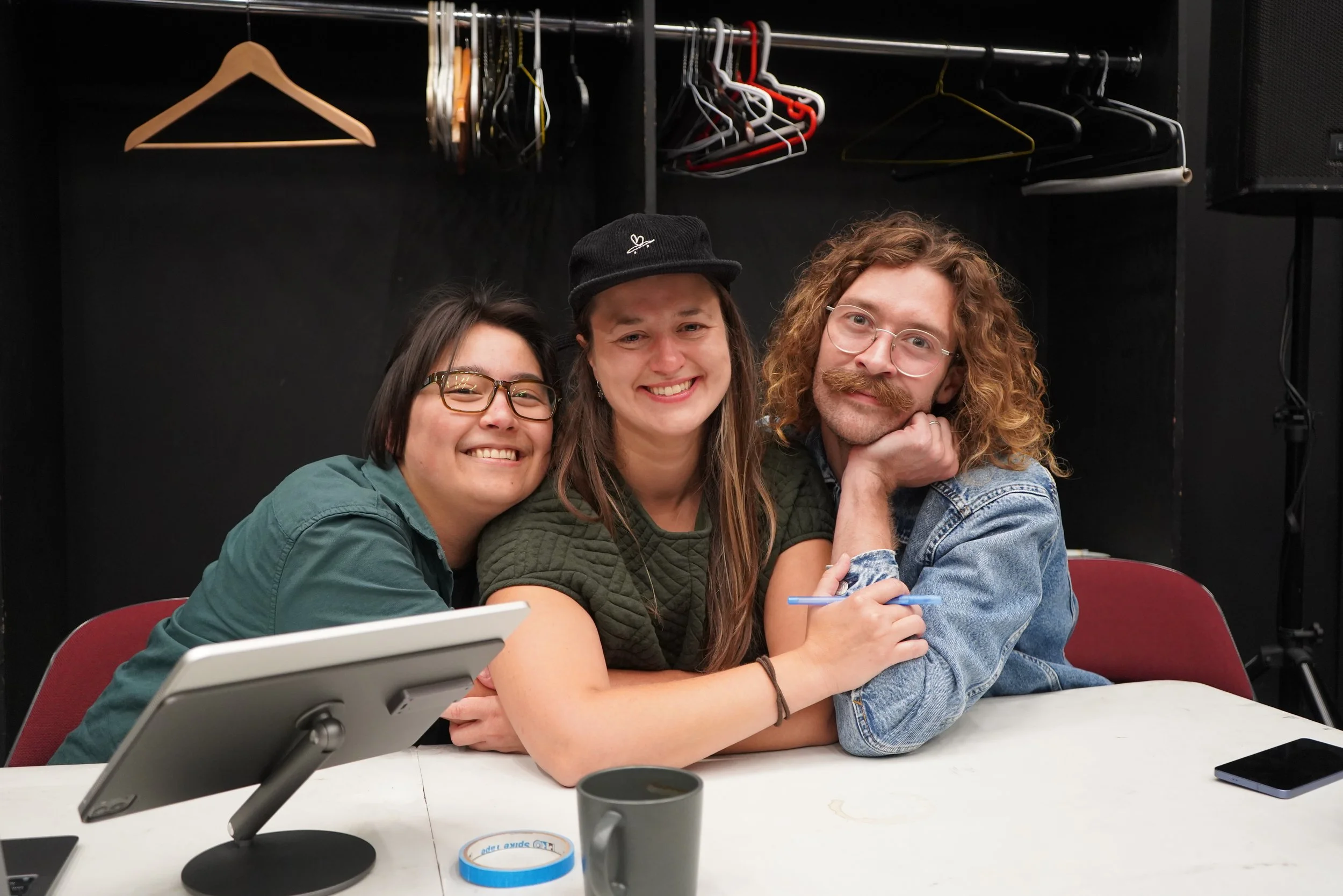 Créations In Vivo. Dillon Orr. Trois personnes souriantes assises à une table, deux femmes et un homme avec des cheveux roux et lunettes, entourées d'objets de bureau, avec un fond noir et un vestiaire visible en arrière-plan.