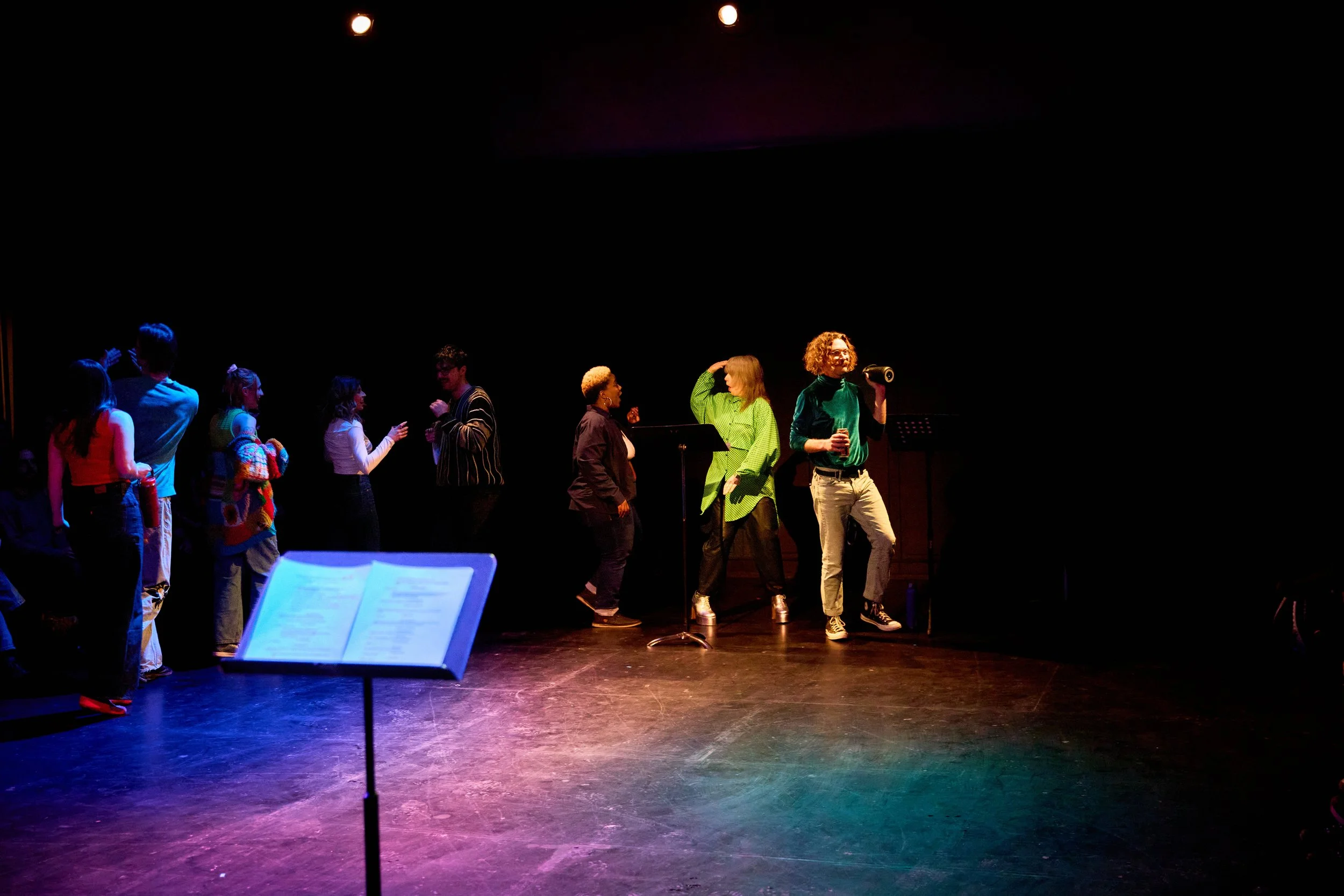 Groupe de personnes en train de discuter et de se détendre sur scène dans une pièce sombre équipée d'éclairage coloré, avec un pupitre à partition au premier plan.