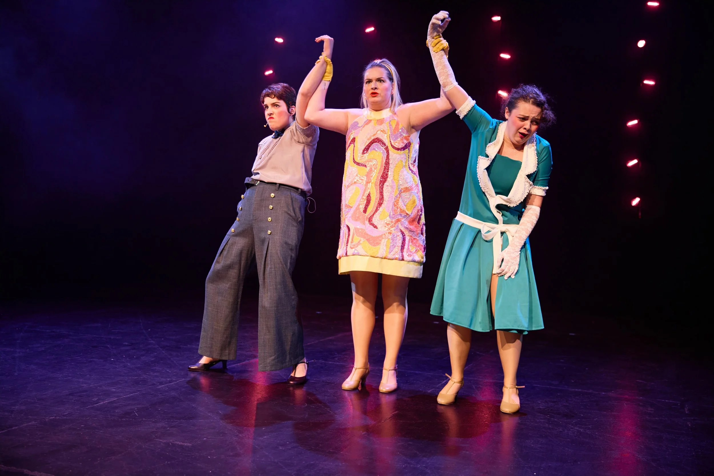 Trois femmes sur scène, debout et en costume, faisant une performance théâtrale ou musicale, avec une lumière rose et un fond noir.