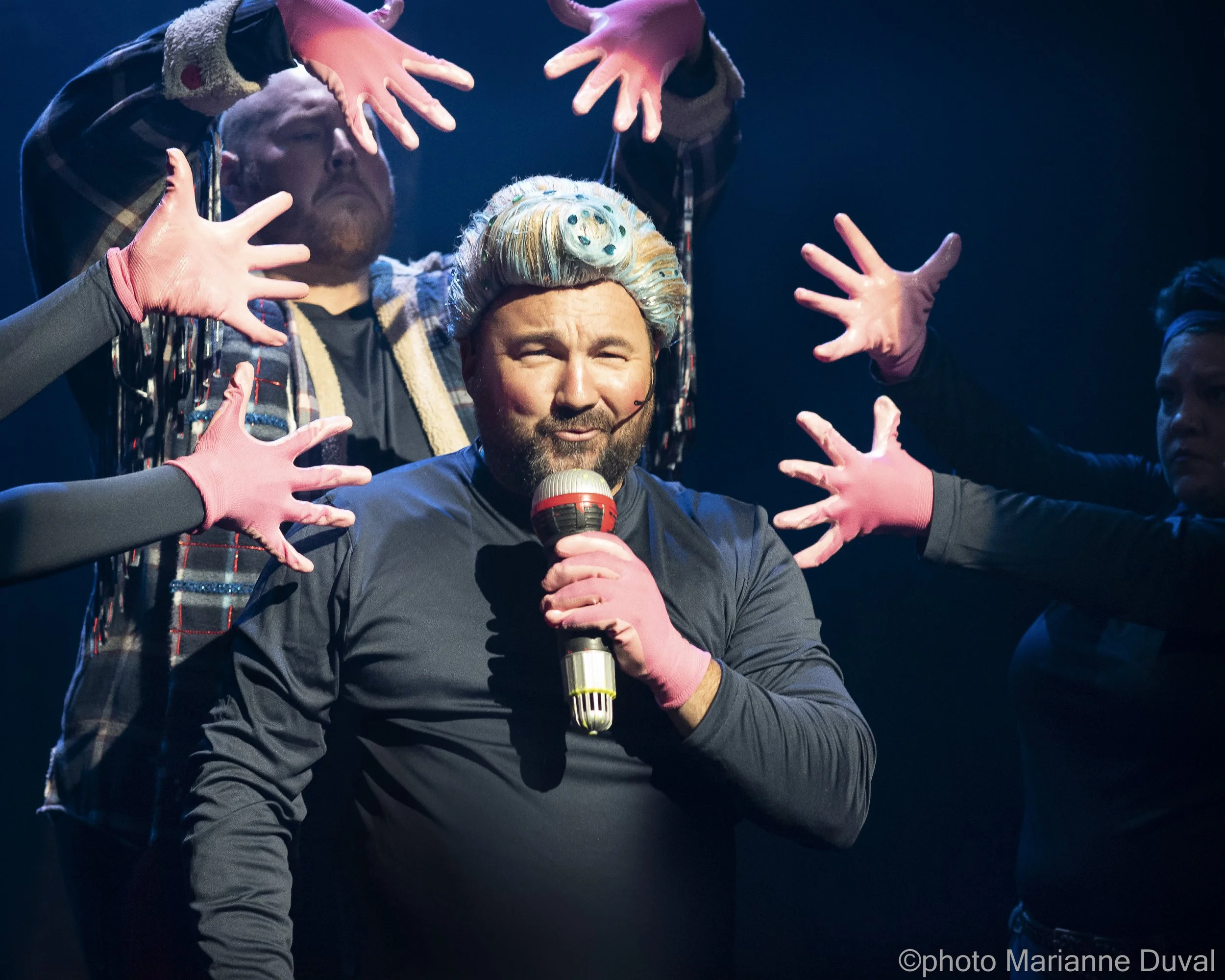 Un homme avec une perruque colorée, tenant un microphone, entouré de plusieurs personnes portant des gants roses, sur scène sous un éclairage sombre.