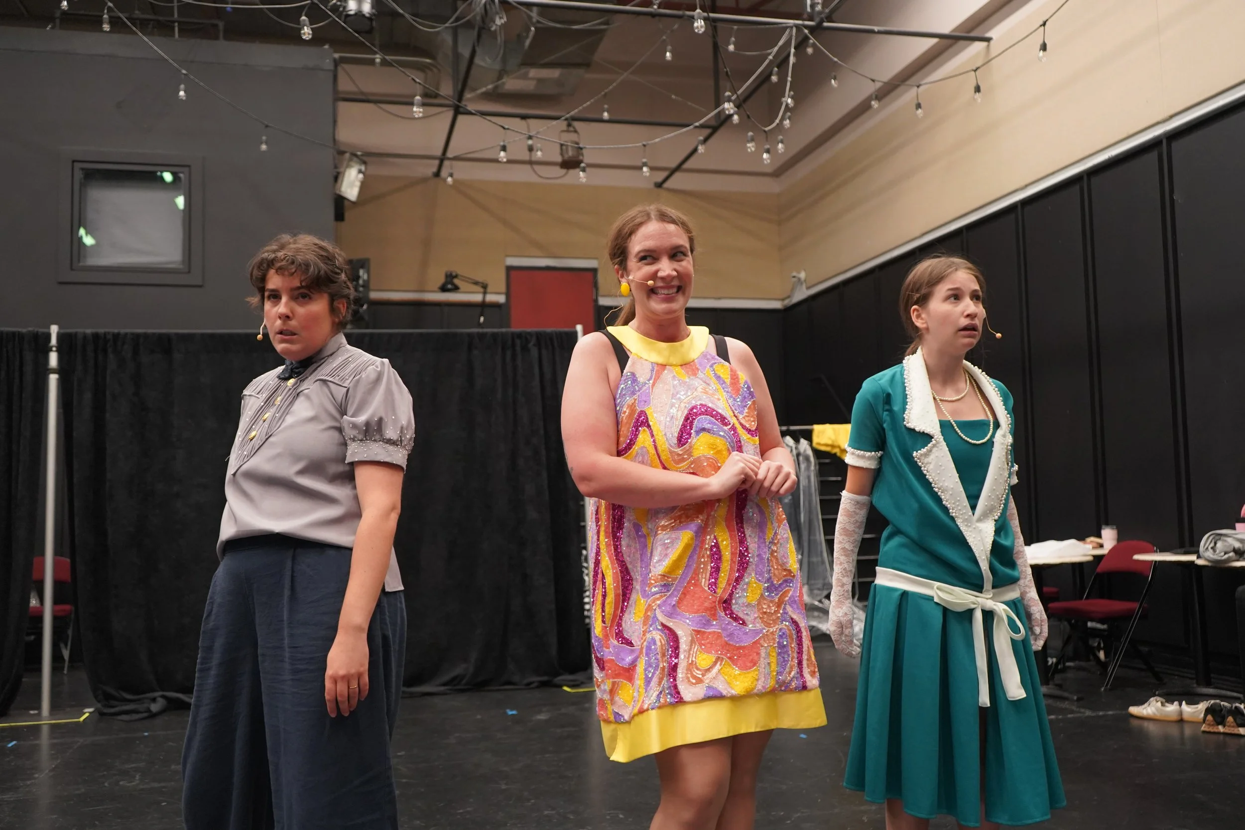Trois femmes debout sur scène, avec des micro-cravates, dans une salle de théâtre ou de répétition, avec un décor simple et des lumières suspendues au plafond.
