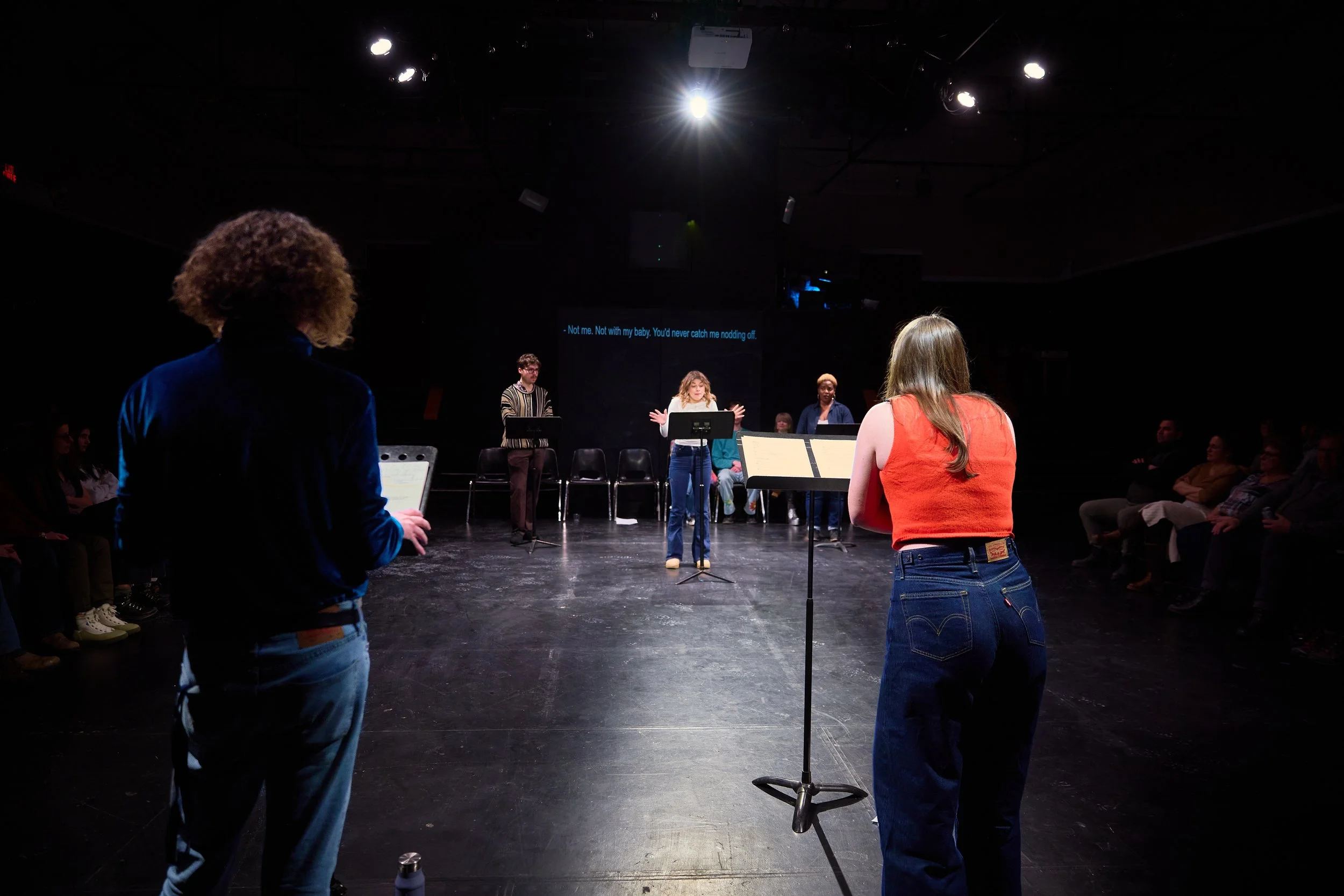 Une scène de théâtre ou de répétition avec huit personnes, dont une femme au centre, qui parle ou chante, entourée de musiciens et avec un public assis dans l'obscurité.