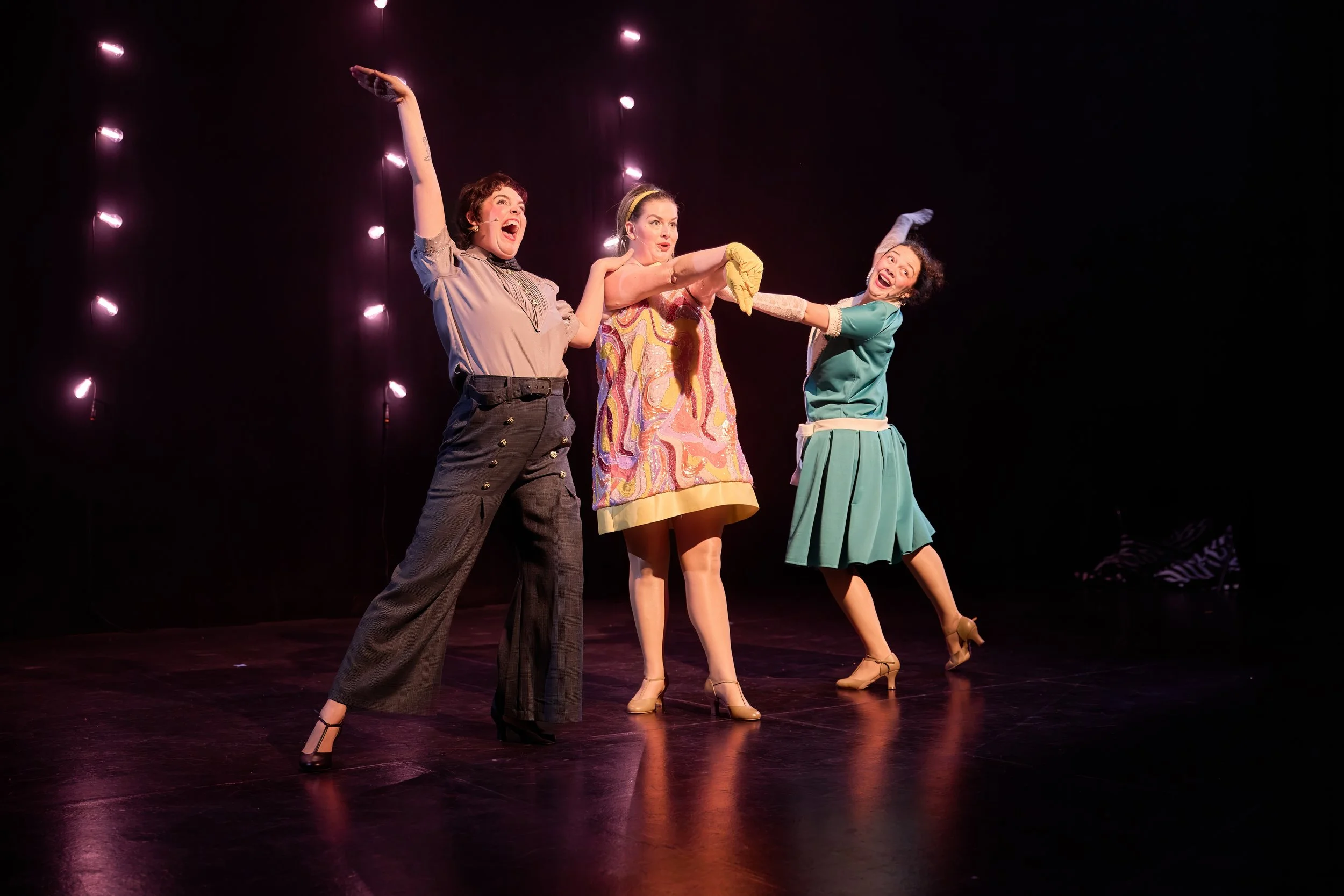 Trois femmes sur scène, en costume vintage coloré, en train de jouer ou de danser, avec des lumières violettes en arrière-plan.