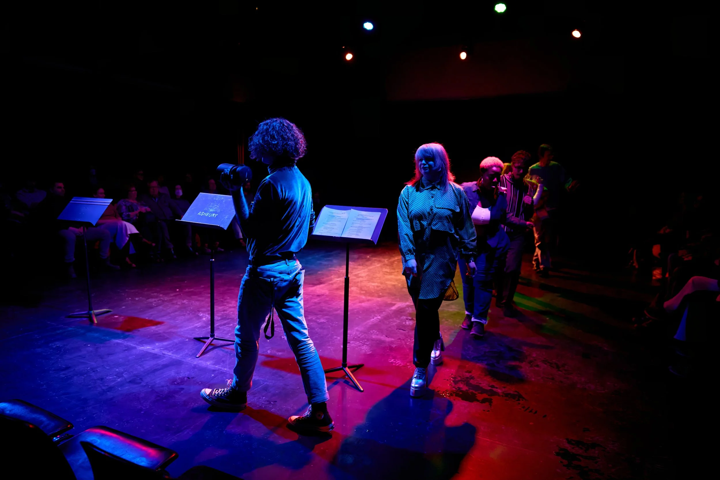 Groupe de personnes sur scène sous des lumières colorées, avec un public assis dans l'obscurité en arrière-plan.
