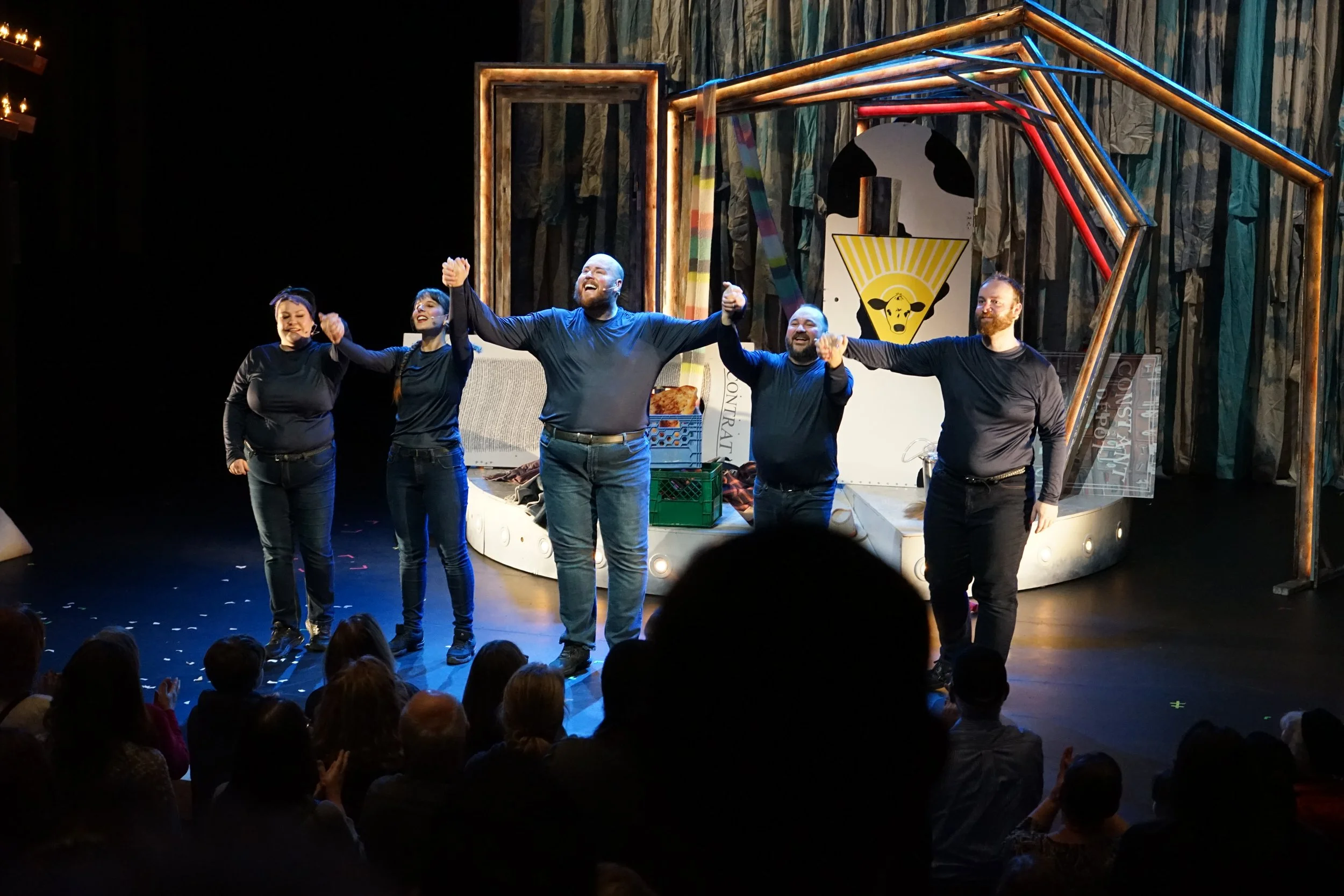 Cinq acteurs de théâtre sur scène, tenant leurs mains levées en signe de victoire ou de fin de représentation, avec un public applaudissant en premier plan, dans un décor théâtral coloré.