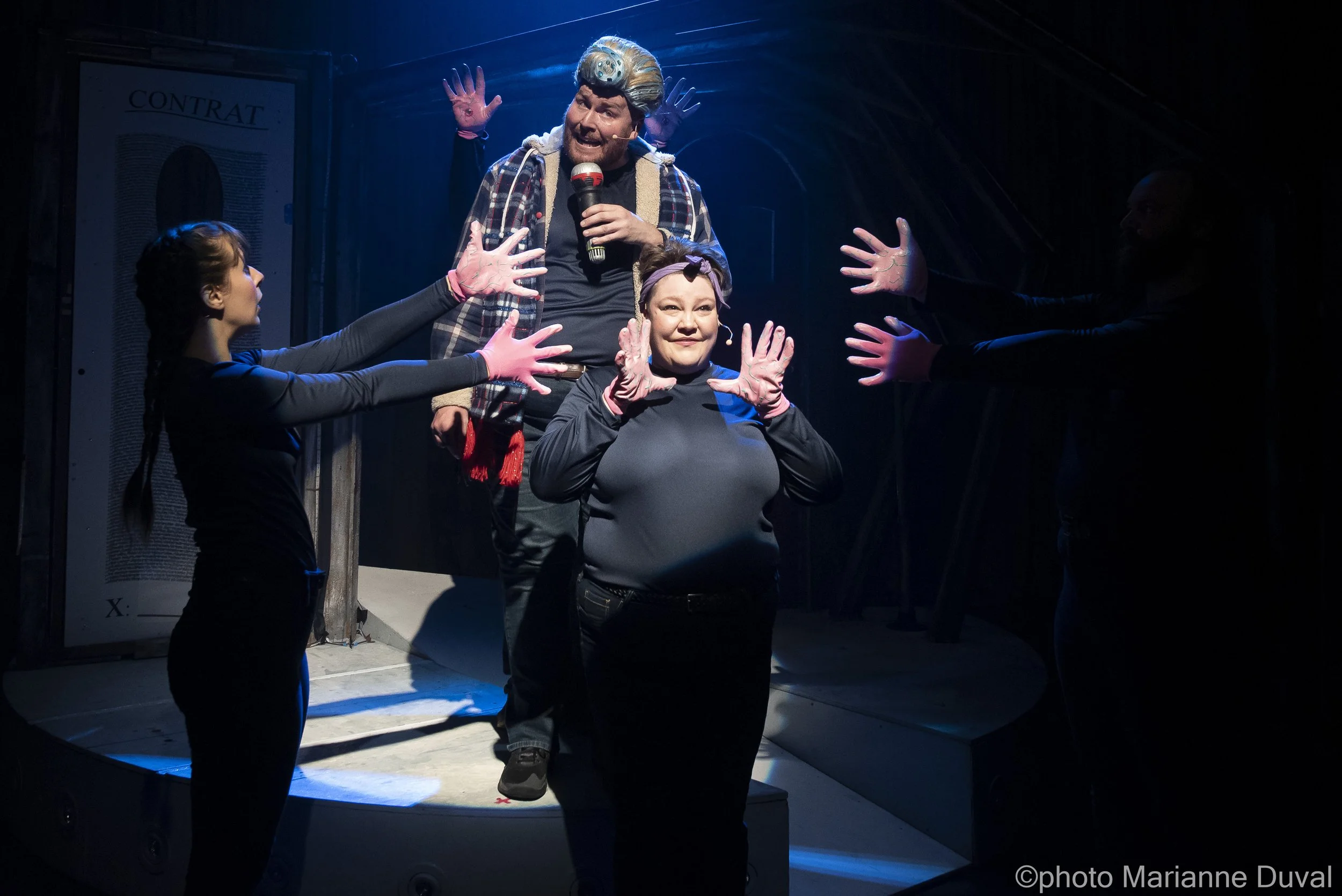 Une scène de théâtre avec deux acteurs, un homme avec une perruque argentée et un micro, et une femme au centre, tous deux entourés de mains gantées. L'homme semble s'adresser au public ou jouer un rôle, tandis que la femme pose pour la caméra. La sc