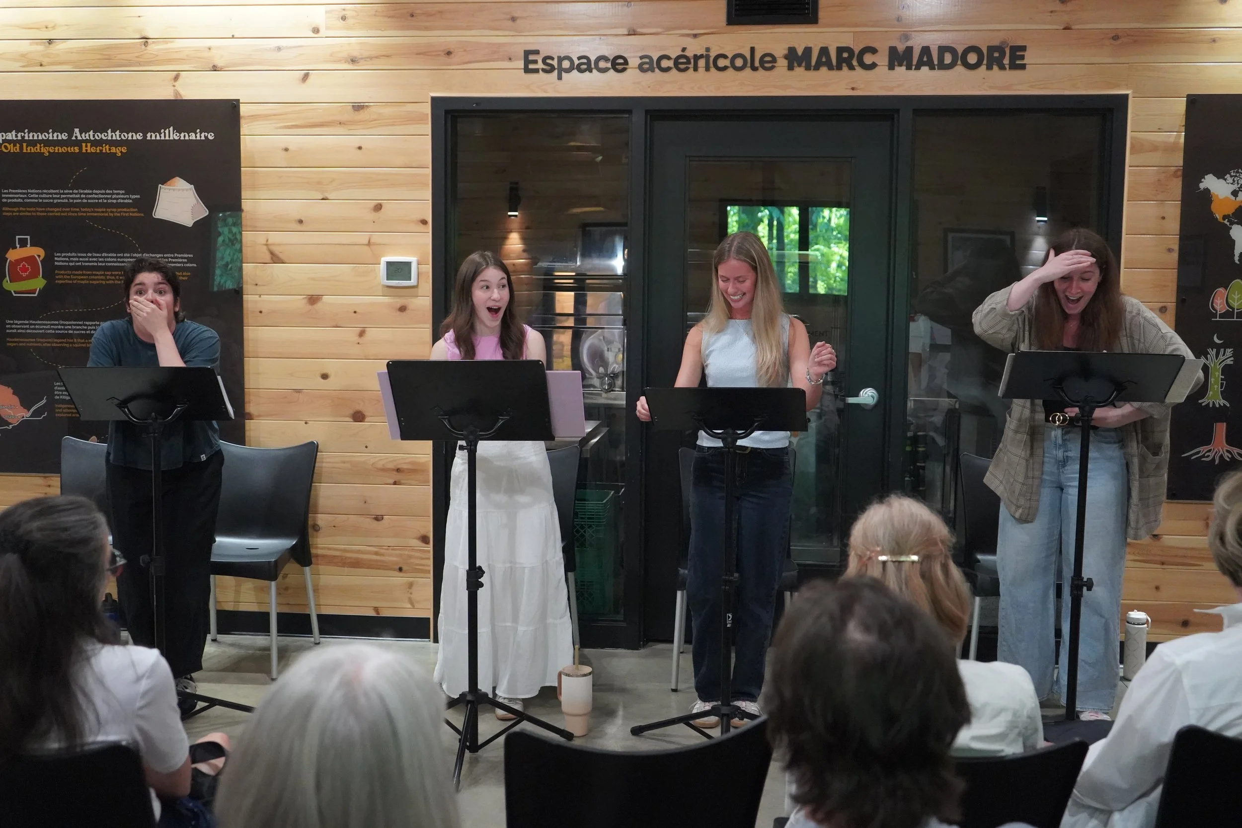 Quatre jeunes femmes se produisent en chantant ou en récitant au microphone lors d'une présentation ou d'un spectacle, avec un public de personnes âgées regardant devant elles.