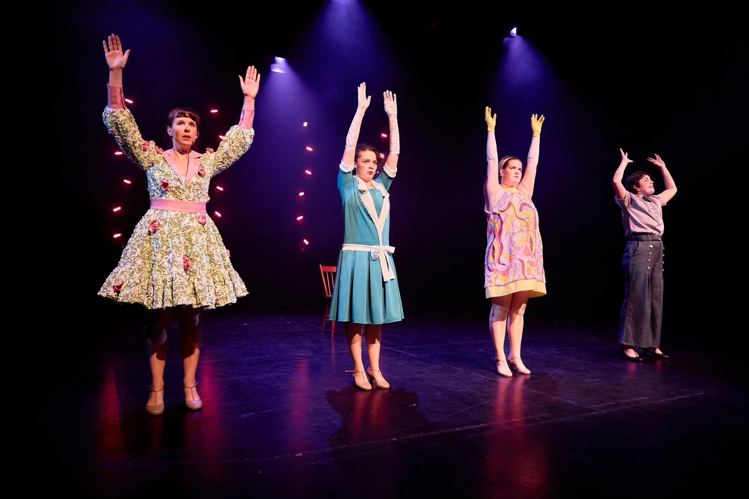 Quatre femmes sur scène en tenue vintage, levant les bras, sous des spots colorés, dans une performance artistique ou théâtrale.