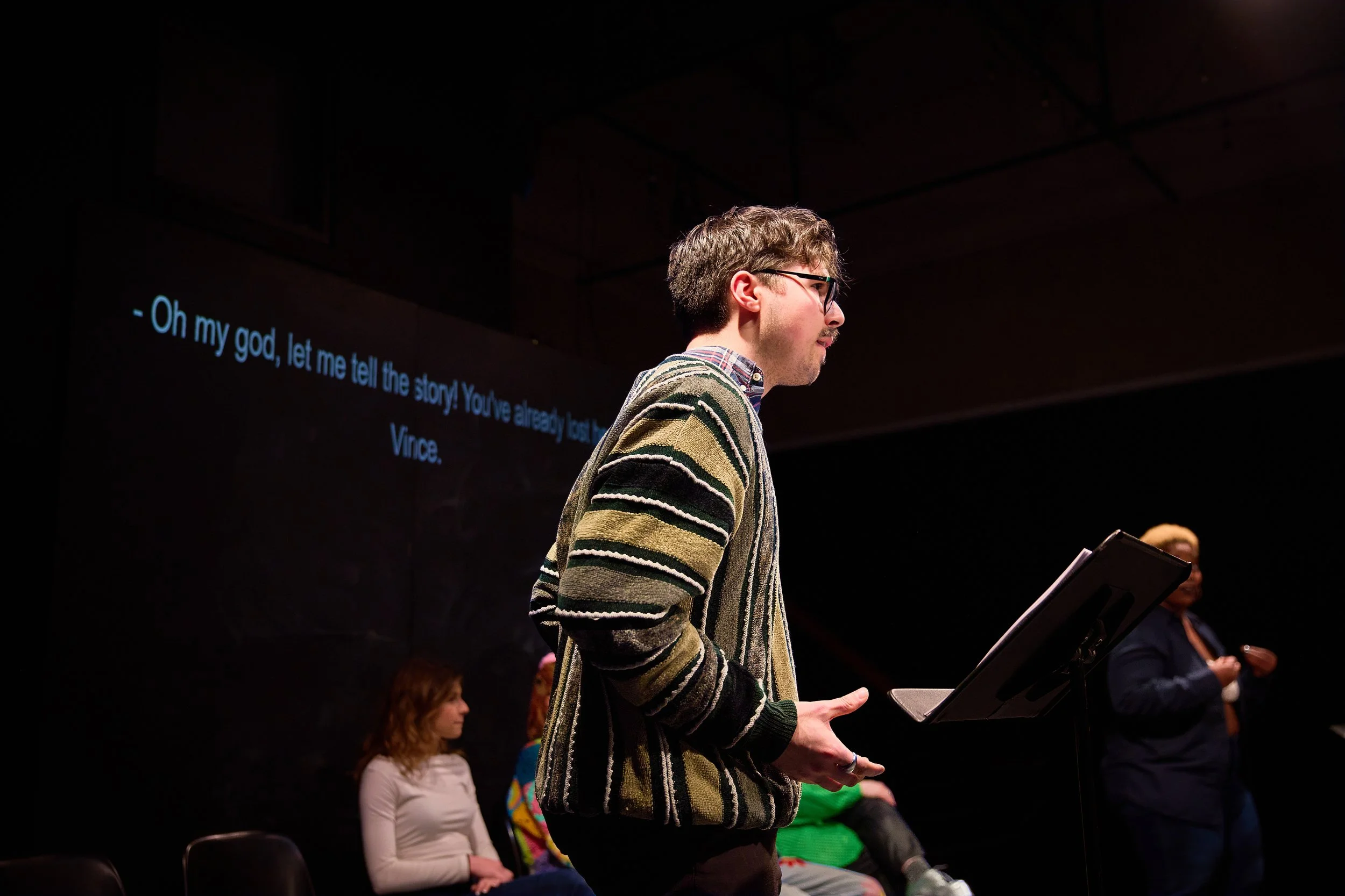 Un homme avec des lunettes, portant un pull rayé, se tient au micro en train de parler lors d'une présentation, avec un écran en arrière-plan affichant un sous-titre en anglais : "Oh my god, let me tell the story! You've already told the story, Vince