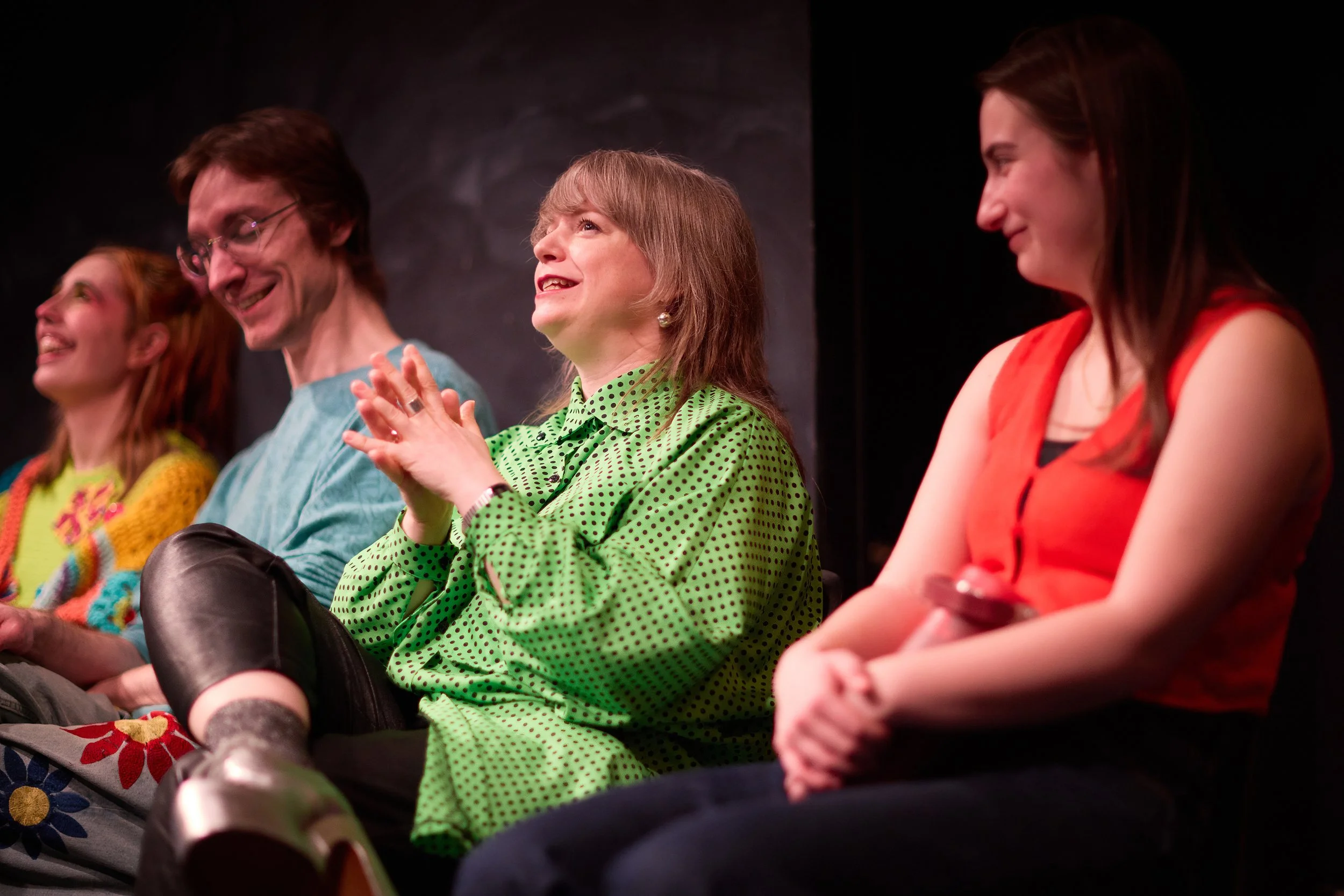 Groupe de quatre personnes assises en formation de spectacle, souriant et applaudissant, dans une scène sombre.