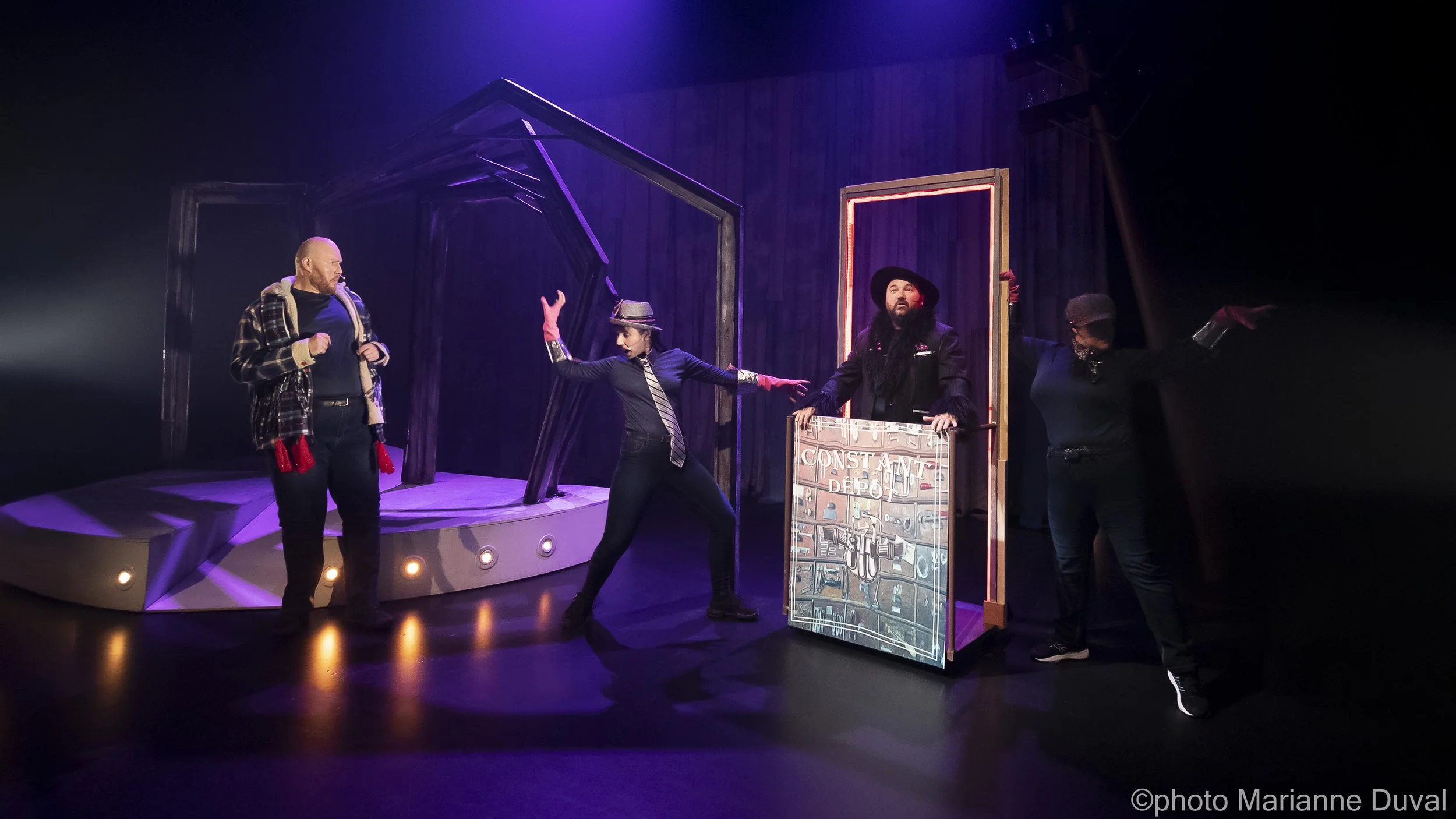 Scène de théâtre avec quatre acteurs. Deux acteurs près du centre, une femme en costume noir avec une veste rayée, un homme avec un chapeau noir. Un homme à gauche porte une veste en flanelle et un pantalon noir. Une personne à droite est en arrière-